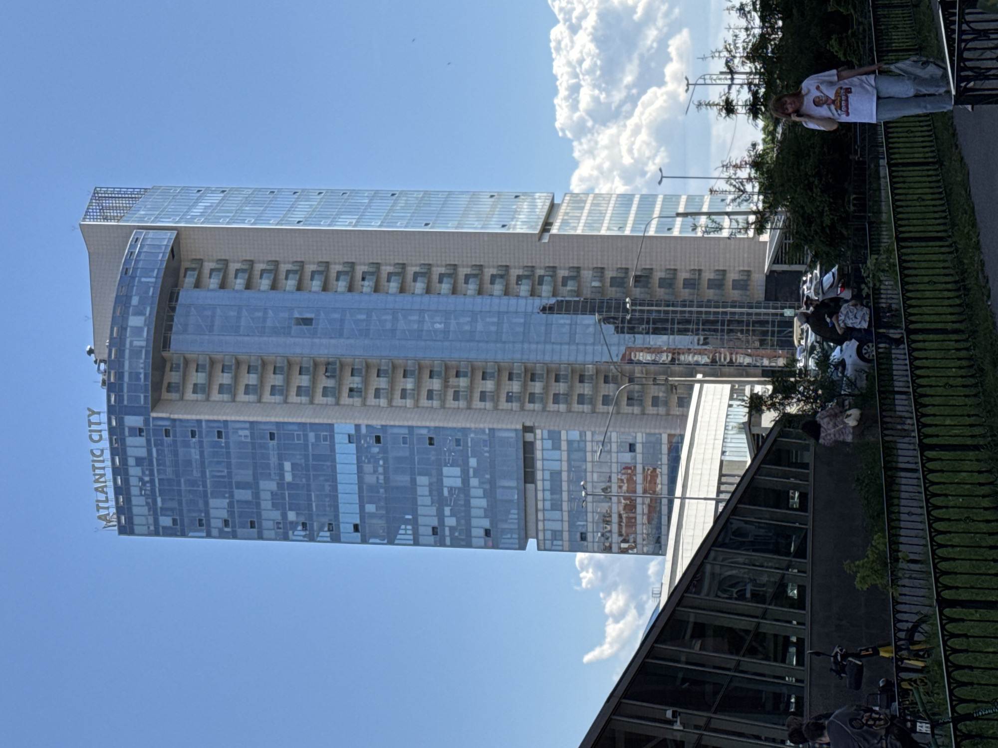 This image depicts a vibrant urban scene in Saint Petersburg, Russia, centered around a modern skyscraper on a clear, sunny day. The dominant feature is a tall, sleek building with a prominent glass facade, displaying the name "ATLANTIC CITY" vertically along its side. This skyscraper features multiple floors with rows of windows and some recessed balconies, topped with a distinctive, multi-paneled glass roof structure.
In the mid-ground and to the right, a black, ornate metal fence separates the viewer's immediate space from a walkway or street. A woman with light brown hair, wearing a white t-shirt with a graphic design and light jeans, stands near this fence, holding a phone to her ear as if in conversation. Beyond her and the fence, there is some greenery and parked cars, with other figures visible in the distance.
In the lower portion of the image, an angular building with a dark, reflective glass roof and facade is partially visible, suggesting a commercial or public space. Several bicycles or scooters are parked nearby, and more people are present in this area.
The time of day is bright daylight, indicated by the clear blue sky and well-lit surroundings. Fluffy white cumulus clouds are visible against the blue. The overall atmosphere is that of a bustling city environment, combining modern architecture with everyday pedestrian and vehicular activity.