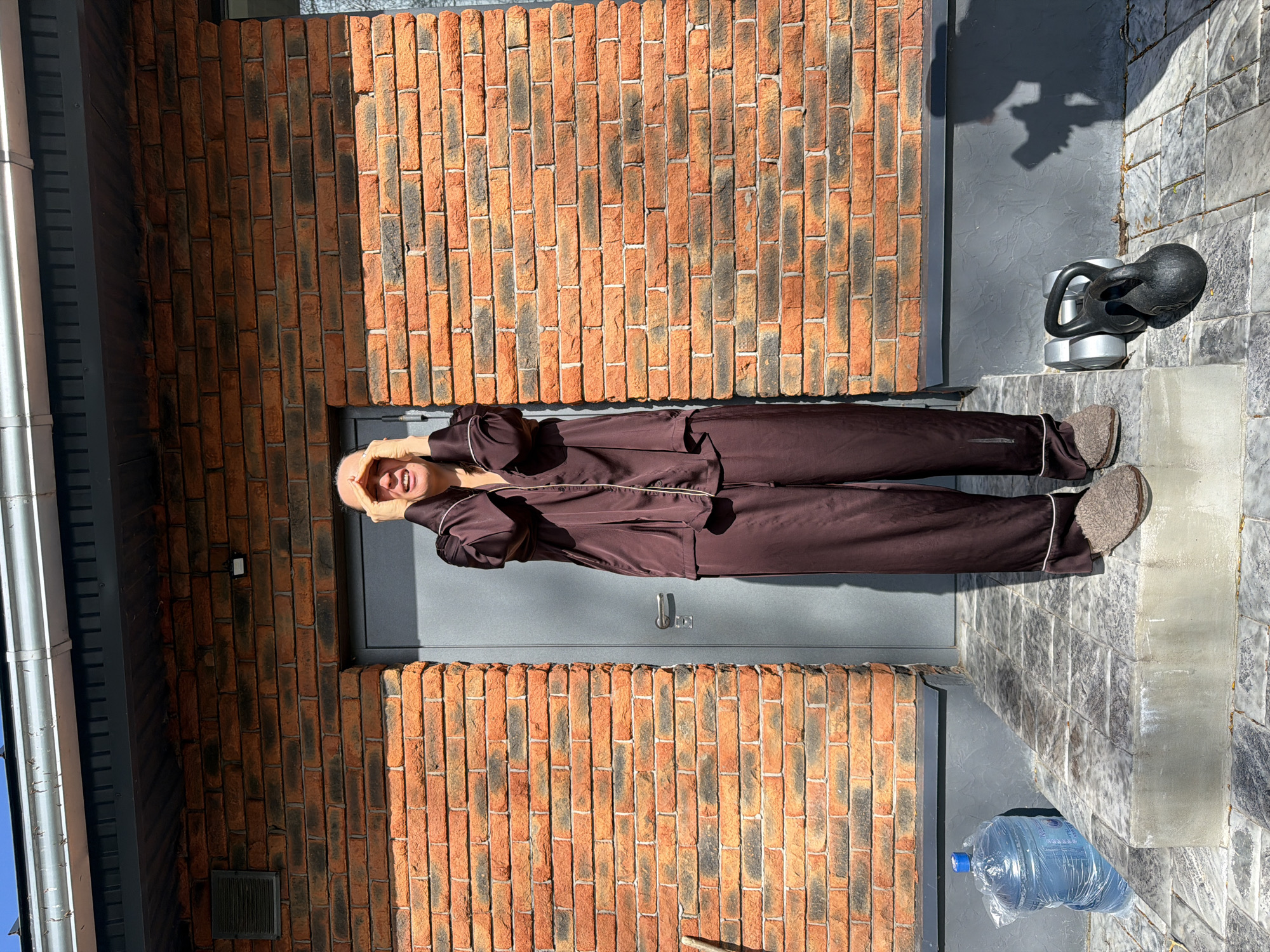 An outdoor, portrait-oriented photograph, taken in Chaikovsky, Russia, captures a person standing casually on a sunny day. The individual is wearing a dark brown pajama set with light piping and light grey slippers, smiling broadly at the camera with hands positioned near their face.
The background features a rustic red and dark brick wall on the left, and a smooth gray-painted wall or door directly behind the person. The ground is a tiled surface in shades of light gray and white. The strong, clear sunlight creates sharp shadows, including the person's shadow on the gray wall and intricate shadows from nearby objects.
To the right of the person, a black kettlebell rests on a pair of silver dumbbells, situated on a slightly elevated platform. Further right and lower in the frame, a large clear plastic water bottle with a blue cap is visible on the ground. Text, likely in Russian, is visible on the water bottle's label. A white drainpipe runs vertically along the far left edge of the scene. The overall impression is one of a cheerful and relaxed moment outdoors.