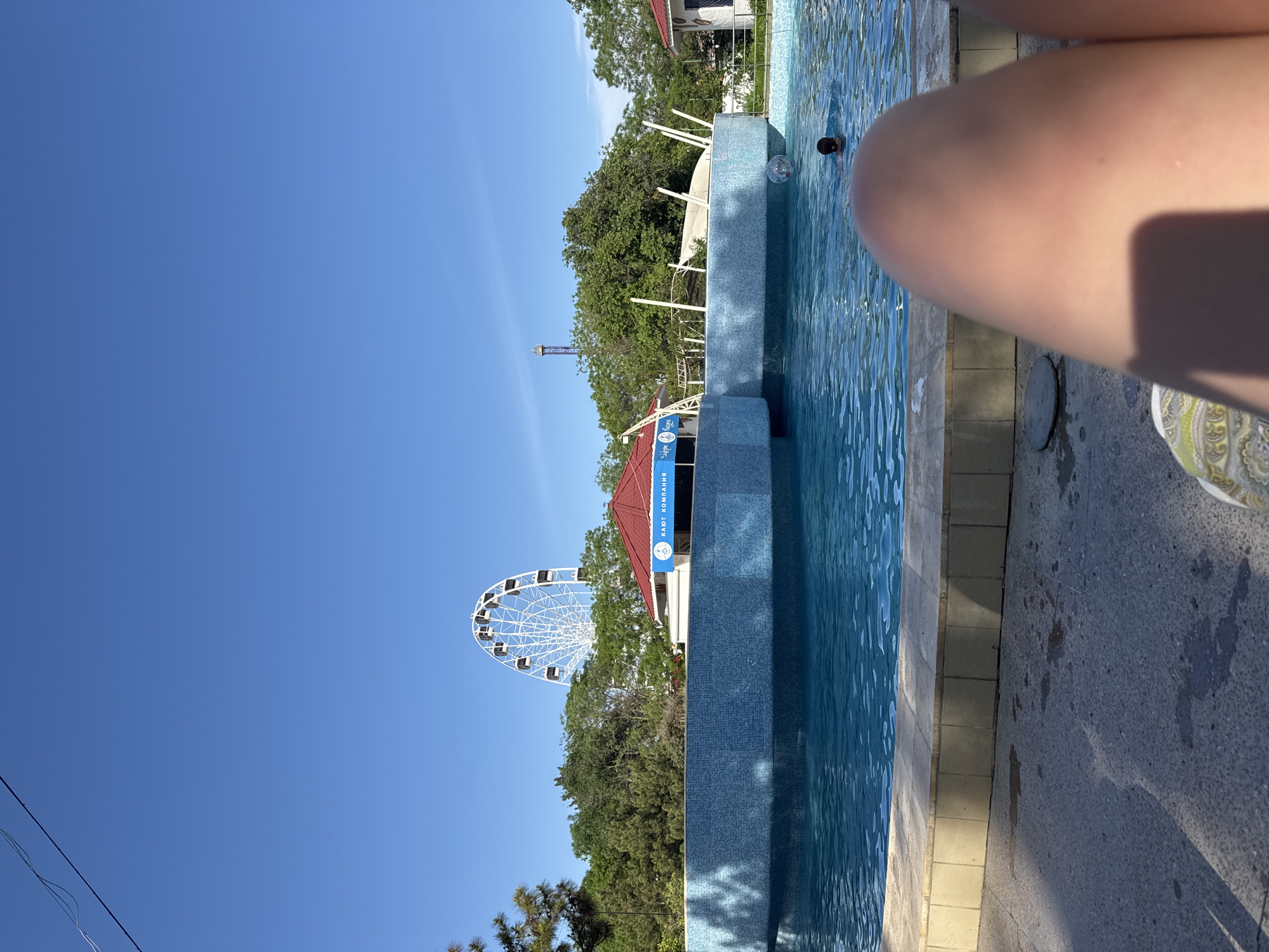 The image, taken in Yevpatoriya, Ukraine, shows a Ferris wheel behind trees, a building, and a swimming pool. A portion of a person's leg is in the foreground on the right. The sky is clear blue.