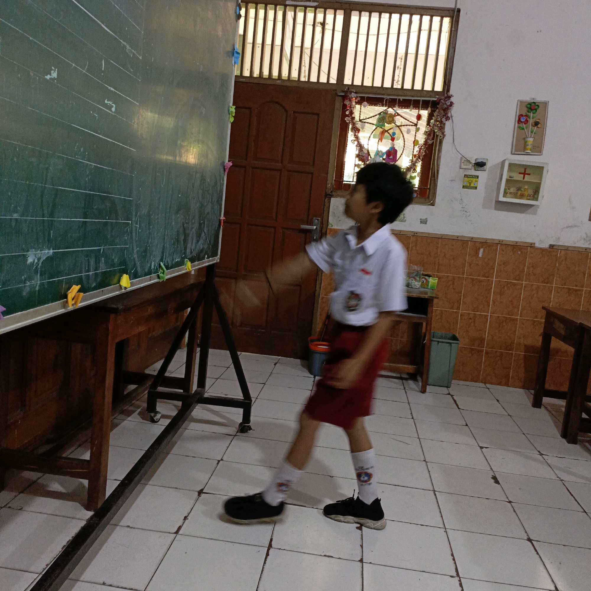 A young boy in a white shirt and red skirt is standing in front of a green chalkboard in a classroom in Salatiga, Indonesia. He is looking up at the chalkboard, and his arm is raised as if he is about to write or point at something. The chalkboard has horizontal lines drawn on it, and some colorful magnets are attached to the bottom edge. The classroom has tiled floors and orange tiled walls. There is a wooden desk in front of the chalkboard, and a wooden door behind the boy. A window with wooden slats is visible above the door. A first aid box is mounted on the wall. The overall scene appears to be a typical classroom setting in a school.