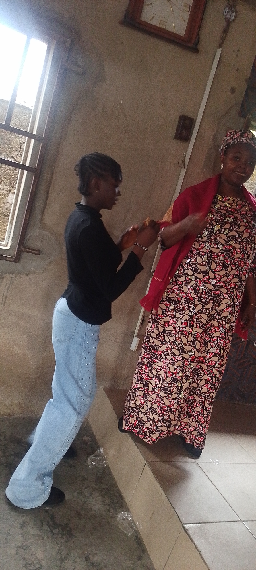 The image depicts two women inside what appears to be a building in Lokoja, Nigeria. The younger woman, wearing a black long-sleeved shirt and light blue jeans with studs, is standing and seems to be adjusting a red shawl or wrap on the older woman. The older woman, who is wearing a patterned, long-sleeved dress and a matching headscarf, is standing on a raised platform with tiled flooring. In the background, there's a window with bars, a clock on the wall, and an electrical outlet. The walls are painted in a neutral color. There are two clear plastic bags/wrappers on the floor.