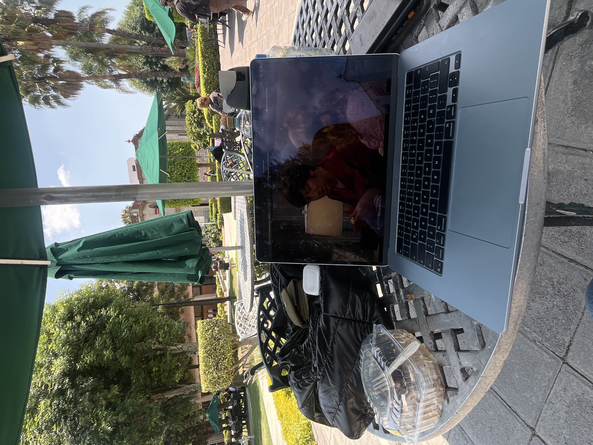 Here is a description of the image:
The image appears to be taken at an outdoor cafe in San Andrés Cholula, Mexico. A laptop is open on a metal lattice-patterned table. The screen of the laptop reflects the light and some of the surrounding area. Near the laptop, there is a packaged pastry and a black jacket. The cafe has green umbrellas. In the background are trees, hedges, and buildings typical of the region. Other people are faintly visible in the background.
