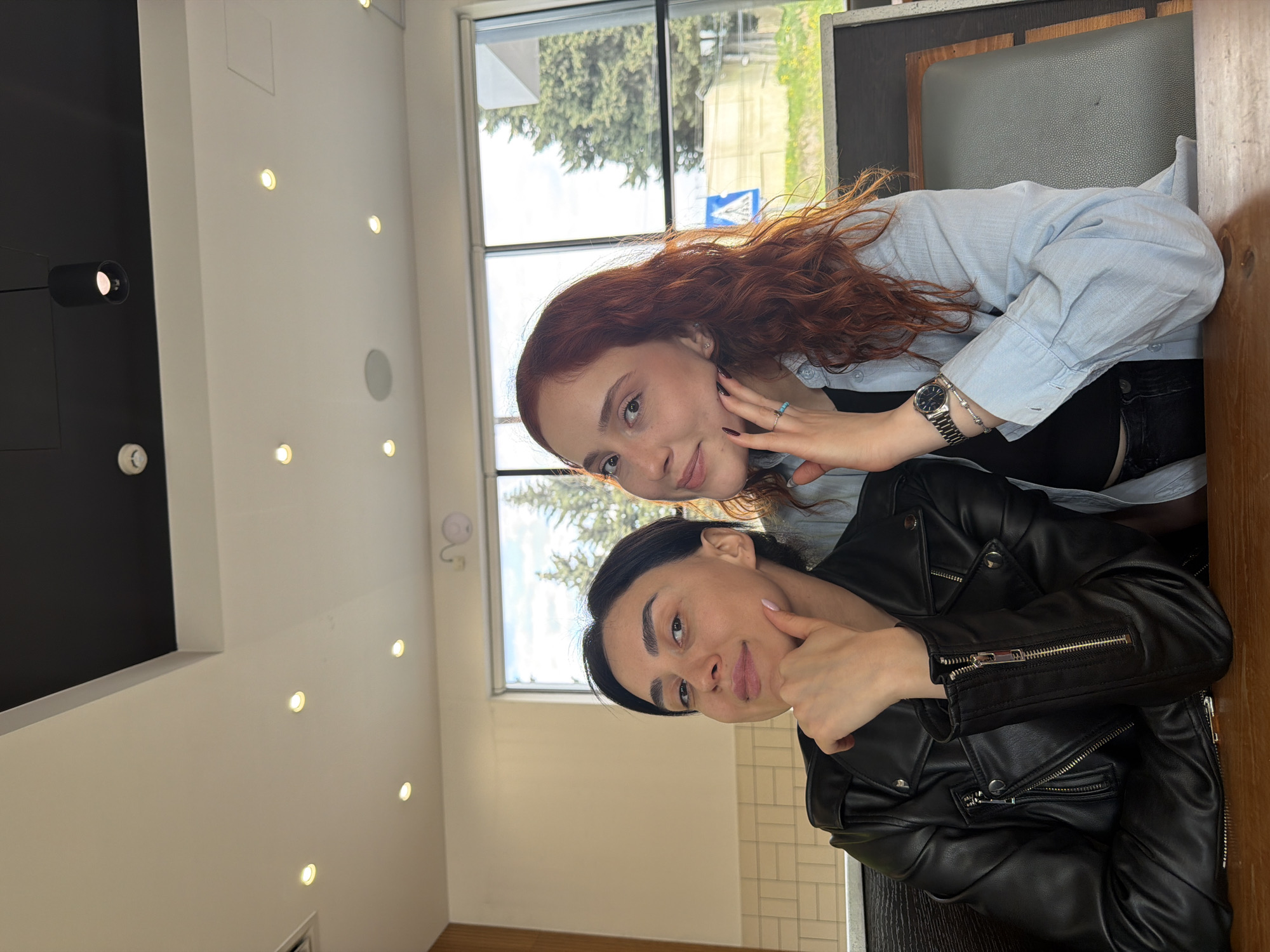 Two young women are captured in a bright, daytime indoor setting, likely a cafe or restaurant in Tbilisi, Georgia. The image is rotated, showing the women's heads pointing towards the top.
The woman in the foreground has dark hair and is wearing a black leather jacket. She smiles subtly at the camera, with her right hand raised, thumb pointing towards her cheek. Behind her, a woman with long, wavy auburn hair also smiles, looking directly at the camera. She is dressed in a light blue long-sleeved shirt over a black top and black pants. She wears a silver watch, a bracelet, and a turquoise-colored ring on her right hand, which she gently places near her face.
The background features large windows revealing a clear sky and green trees outside, along with a blue road sign. The interior has white walls with multiple recessed spotlights on the ceiling, creating a warm ambiance. A section of the ceiling or wall is painted black, featuring a dark spotlight fixture. Booth seating with upholstered backs and wooden accents is visible, suggesting a comfortable dining atmosphere. Both women appear relaxed and cheerful. No discernible text is visible other than the distant road sign.