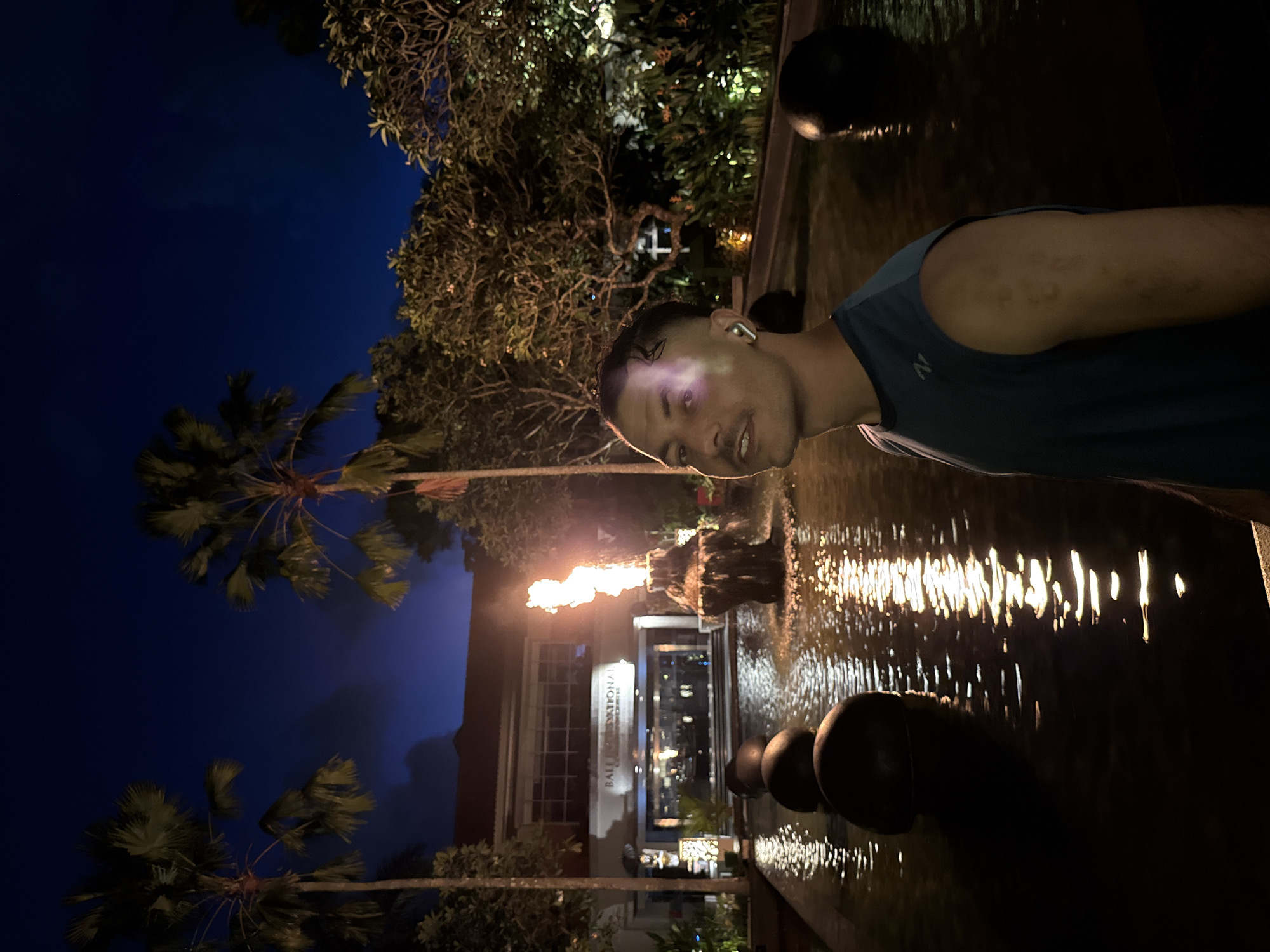 The photograph captures a man wearing a blue tank top standing alongside a decorative water feature at night. He has an earpiece in his left ear. The water feature consists of a dark, reflective pool, with multiple dark spherical elements partially submerged. In the background, flames rise from a water fountain, illuminating a building with the inscription "Bali International" on the facade. Palm trees are visible on the left side of the image, silhouetted against the dark blue sky. The location is in the city of Unknown, Indonesia.
