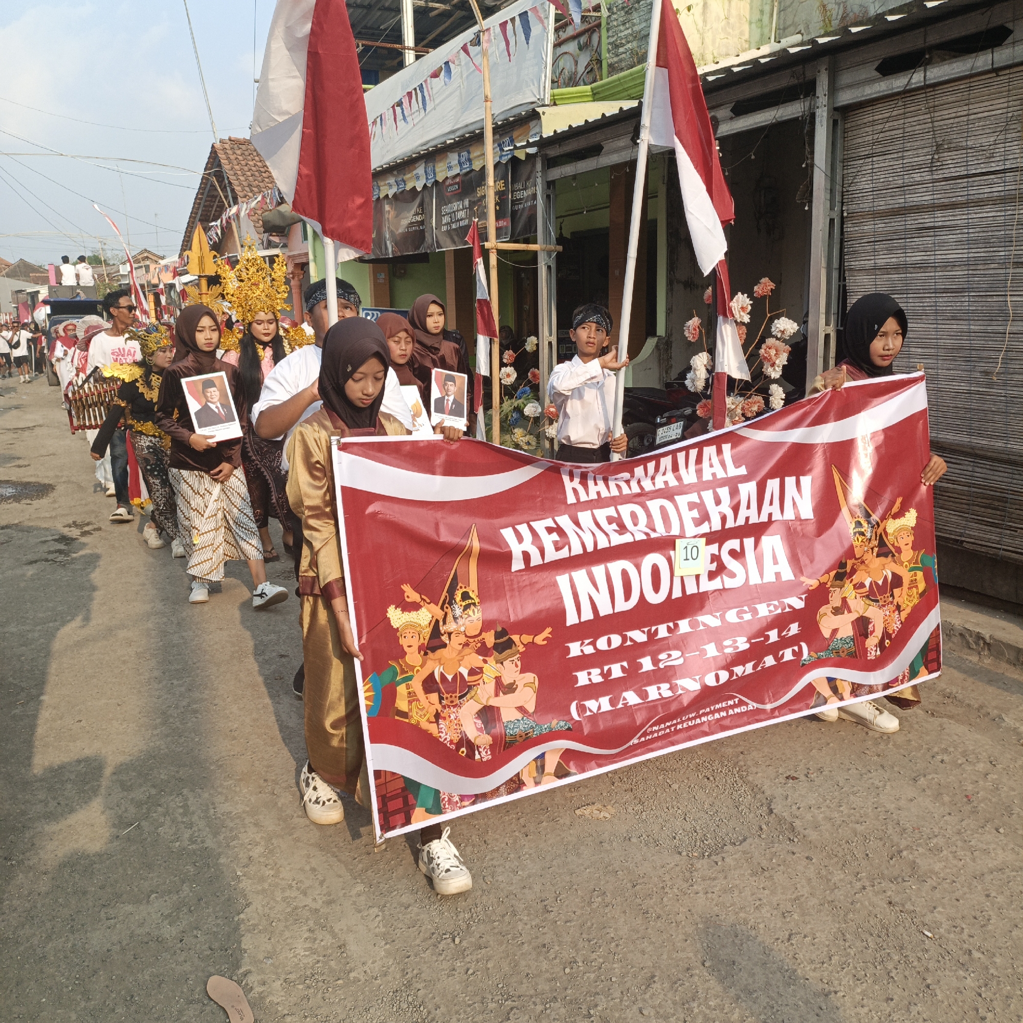 In Tanahbaya, Indonesia, a procession is underway on a street lined with buildings. A large red banner is held horizontally by several people. The banner reads "KARNAVAL KEMERDEKAAN INDONESIA" (Indonesian Independence Carnival) and includes the text "KONTINGEN RT 12-13-14 (MARNOMAT)". Below this, smaller text is partially visible, reading "@NANAUW.PAYMENT SAHABAT KEUANGAN ANDA". The banner is decorated with depictions of traditional Indonesian dancers. In front of the banner, several individuals, primarily young women, are walking. One woman in the foreground wears a gold-colored outfit and a headscarf, while another in a brown headscarf and traditional patterned skirt holds a framed portrait of a man. Behind them, other participants are visible, some holding Indonesian flags. The street appears to be paved with asphalt and gravel. The sky is clear and a warm sunlight illuminates the scene, casting shadows on the ground. Buildings with tiled roofs and corrugated metal facades are visible on either side of the street.
