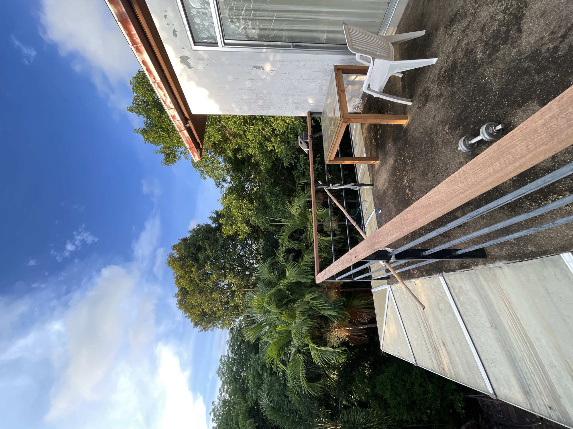 The image depicts an outdoor area, likely a balcony or patio, in Baan Thong Po, Thailand. A section of a building with a window is visible, along with a white plastic chair and a glass-topped table with a wooden frame. A single dumbbell lies on the concrete surface of the patio. A railing with horizontal metal bars and a wooden top runs along the edge. Lush green foliage, including trees and palm trees, extends beyond the railing. The sky is blue with scattered clouds.