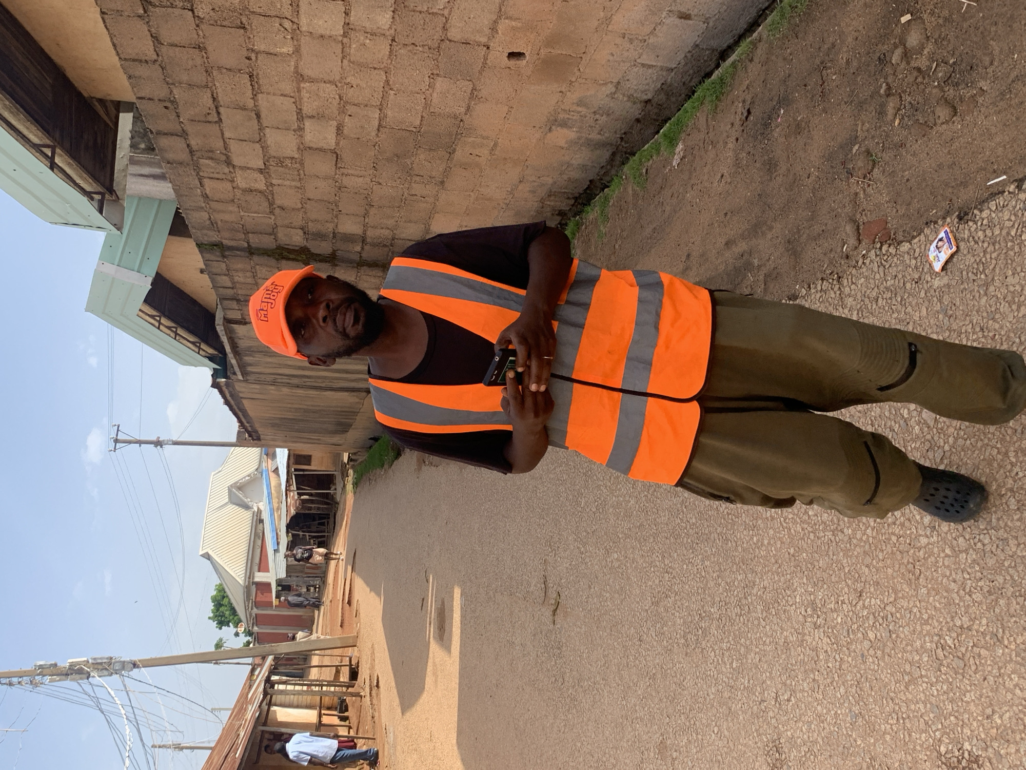 A man wearing an orange safety vest over a black shirt and dark pants stands in what appears to be a street in Jos, Nigeria. He also wears an orange baseball cap. The man is holding a black mobile phone in his right hand. The background consists of a brick wall on the left and a dirt pathway with some vegetation on the right. Further in the background, there are buildings and a street. The sky is visible above, suggesting it is daytime, likely a sunny day. The overall scene is that of a street in a residential or commercial area. The man's expression is neutral. There are no other people in immediate proximity to the man, though some figures are visible further down the street. The image is oriented sideways.