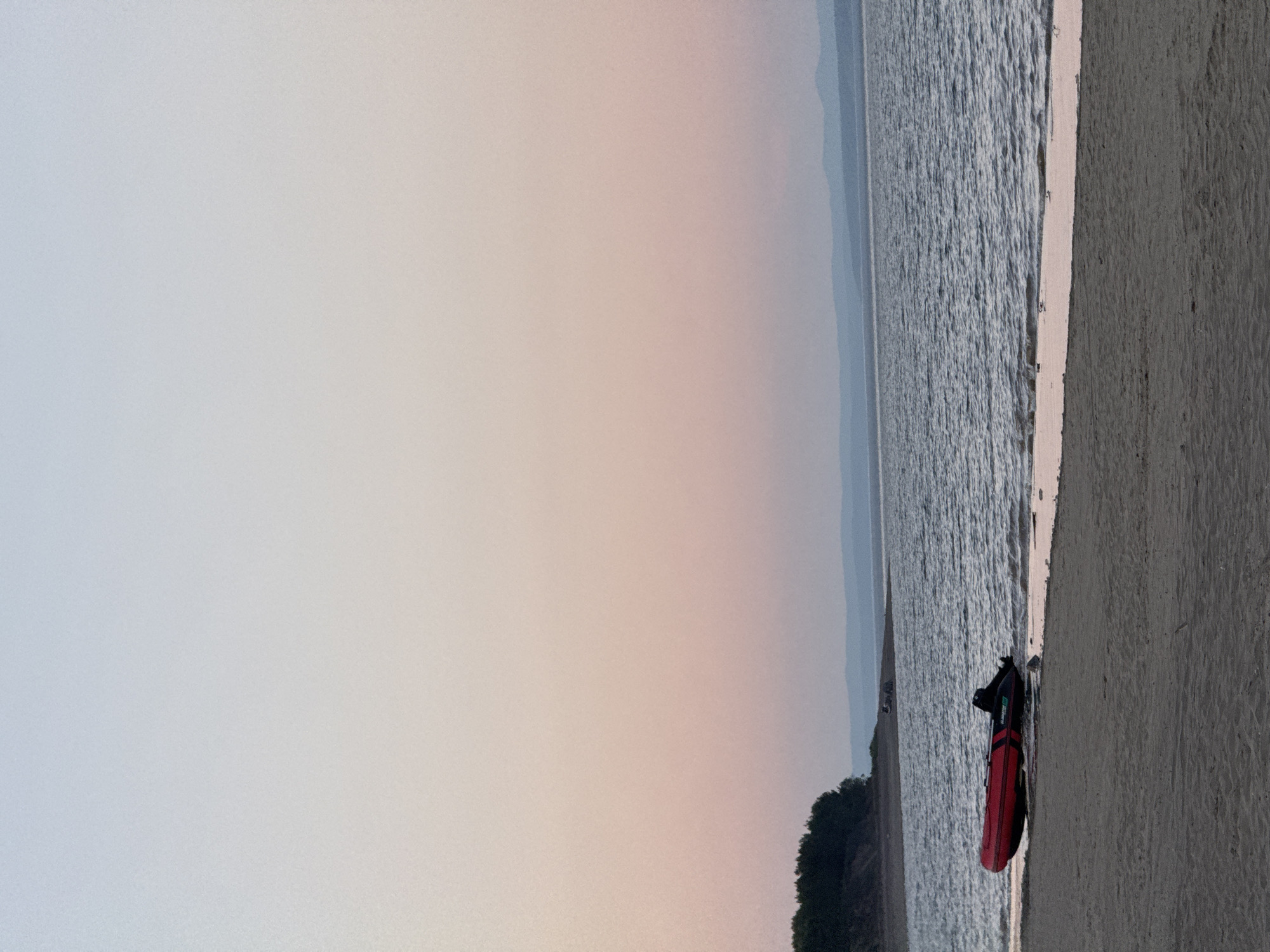 This image captures a panoramic view of a large body of water and a sandy beach in Daurskoe, Russia, during twilight. The sky displays a clear gradient, transitioning from a light blue at the top to a soft pink and peach near the horizon. In the distance, faint, layered silhouettes of dark blue mountains or hills are visible along the horizon line. The water surface shows gentle ripples, subtly reflecting the sky's colors. On the far left, a dense cluster of dark green trees forms a forested edge on a raised bank. A red inflatable boat, labeled "GLADIATOR" on its side, with an attached outboard motor, is partially beached on the sandy shore. The boat's bow faces left, with its stern and motor partially submerged in the water. The foreground is dominated by a wide expanse of light brown, undisturbed sand. No individuals are visible in the scene.