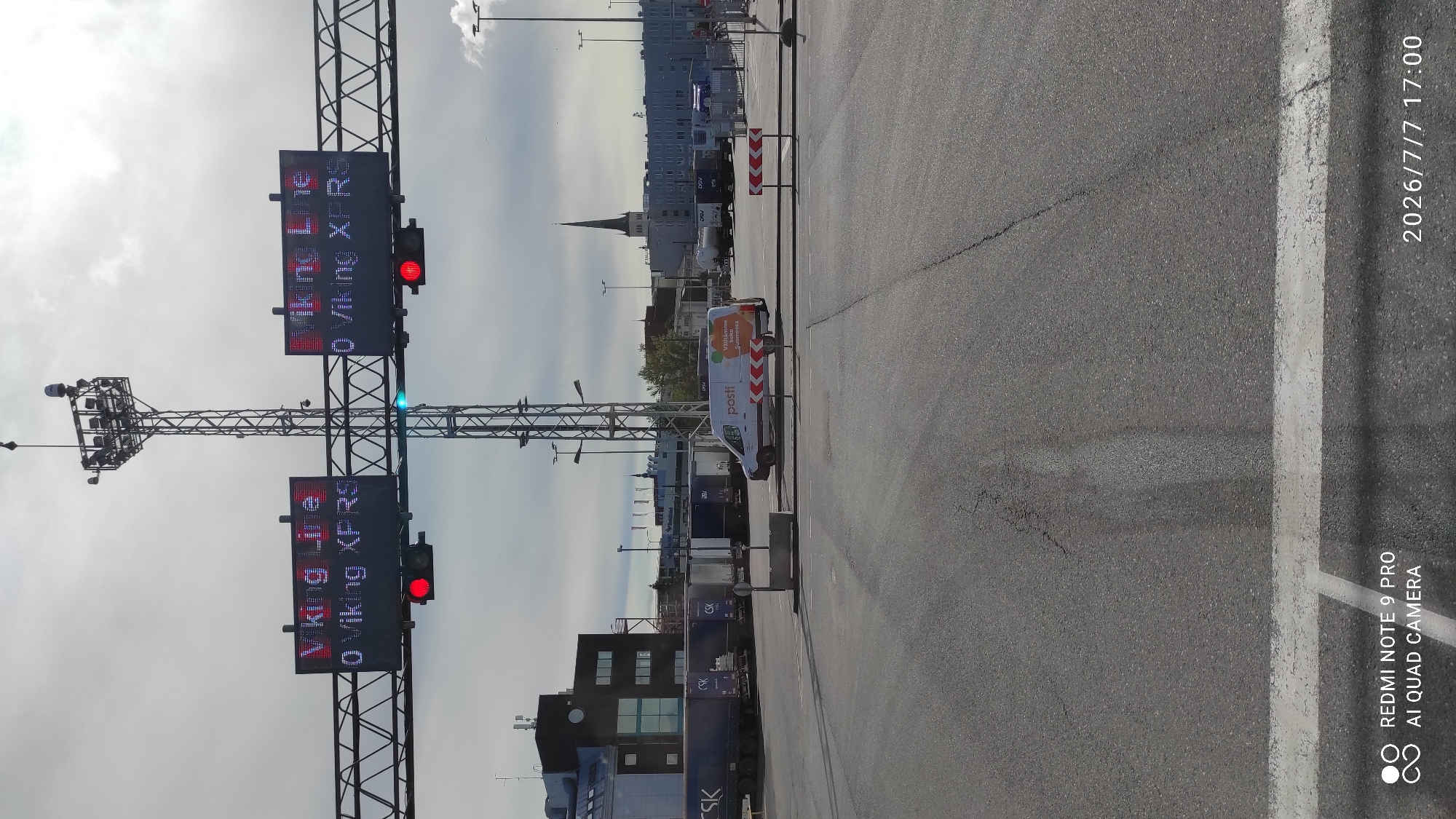 An overhead shot captures a ferry terminal entrance in Tallinn, Estonia, under a cloudy sky. Two large digital signs display "VIKING LINE" in red and "0 VIKING XPRS" in white, with red lights illuminated below them, indicating a potential stop. The terminal area is paved with asphalt marked with white lines. Several trucks and a white Posti van are visible in the background, along with a tall tower and a building with a spire. The image also includes timestamps in the bottom right corner.