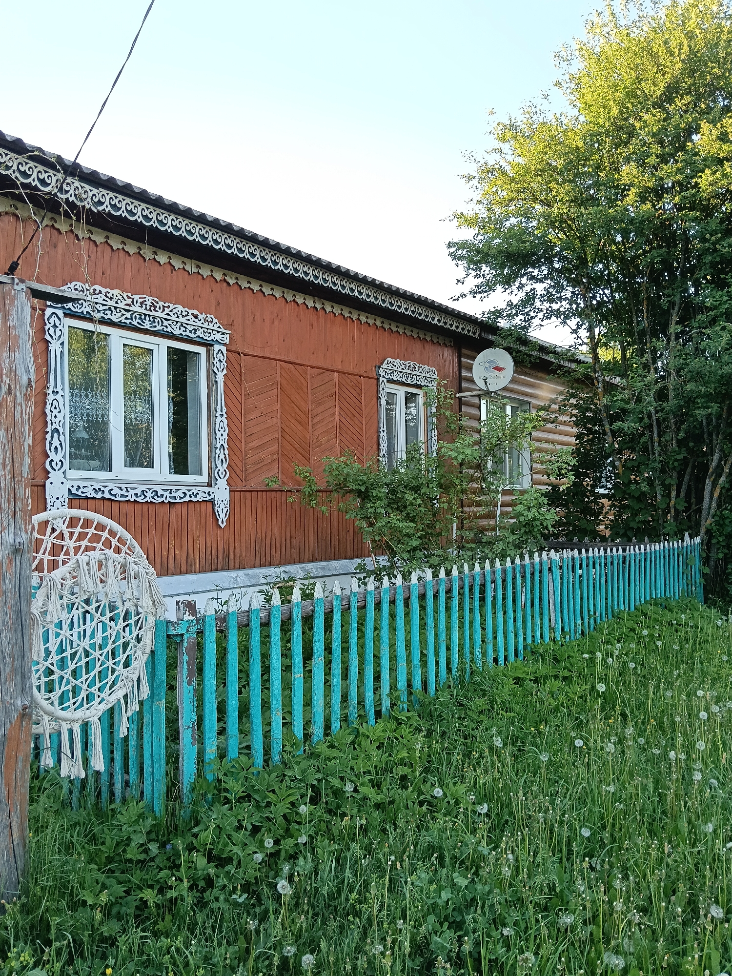 A residential scene shows a single-story wooden house with ornate, white trim around the windows and roofline, located in Красный Маяк, Russia. The house is painted a reddish-brown color, and there is a satellite dish mounted on the side facing the camera. A turquoise wooden picket fence runs along the front of the property, partially obscured by overgrown vegetation and dandelions. A macrame-style hanging is attached to a wooden post in the foreground.