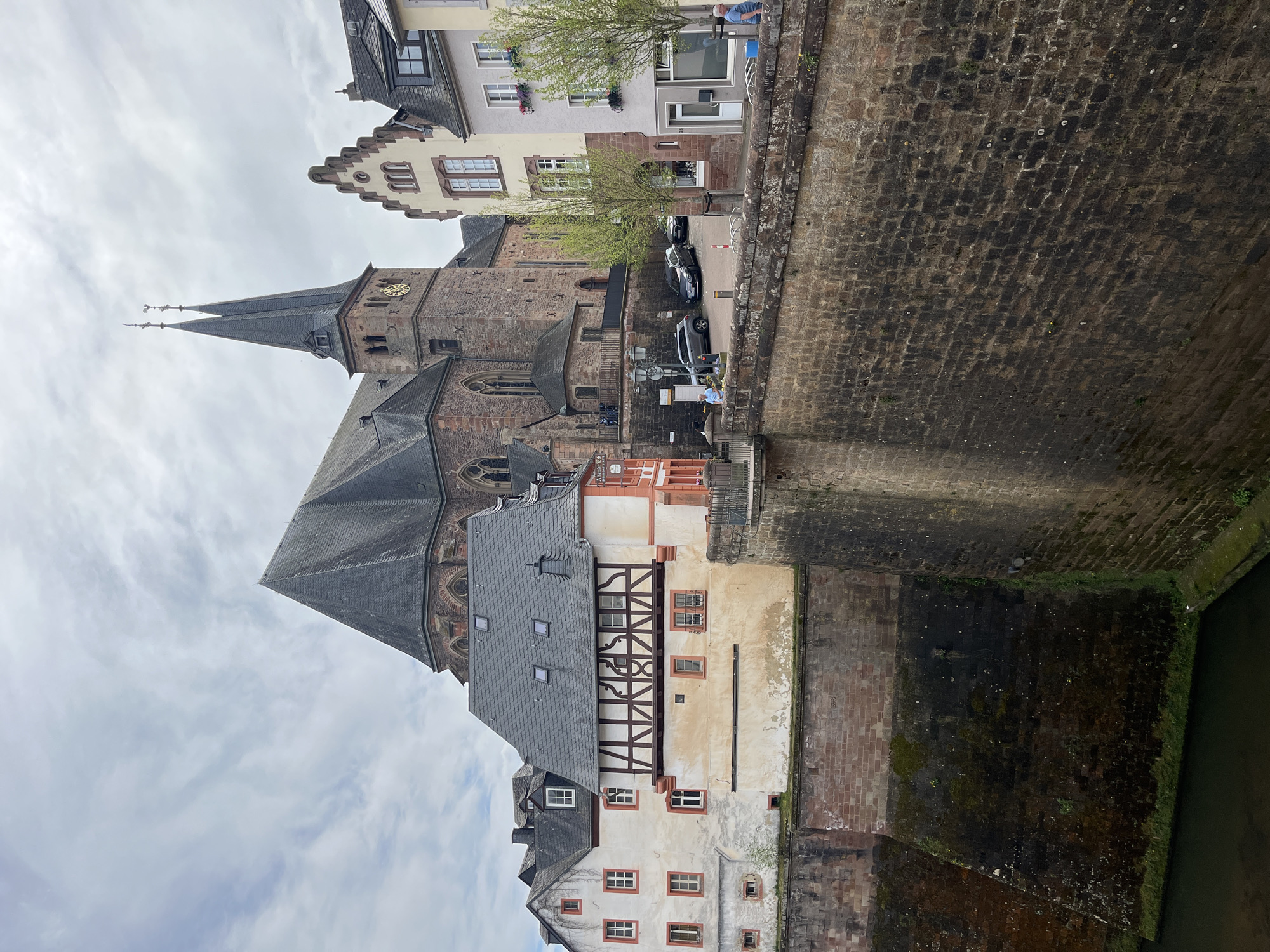 The image shows a streetscape in Saarburg, Germany. A tall, dark-colored stone church with a steeple is prominent. Adjacent to the church are several older buildings with a combination of white stucco and half-timbered facades. A stone wall, possibly part of the riverbank of the Leukbach, runs along the edge of the street. Cars are parked in the street. There are people walking on the other side of the road. The weather is overcast.