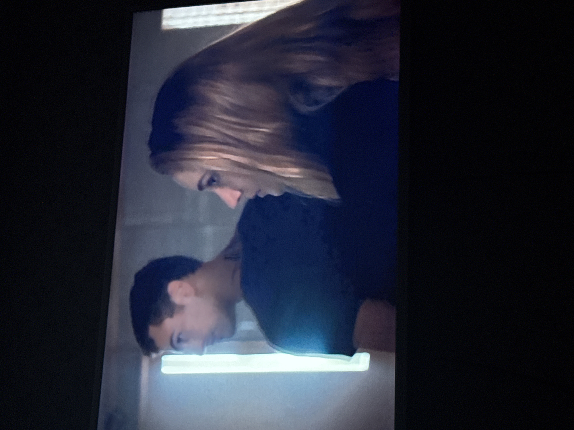 A man and a woman are looking down at an illuminated surface. The man has short, dark hair and is wearing a dark shirt. The woman has long, blonde hair and is also wearing a dark shirt. They are both leaning forward and appear to be examining the surface closely. The surface is white and emits a bright, glowing light. The background is blurry and indistinct. There is no indication this image was taken in the city of Mytishchi, Russia.