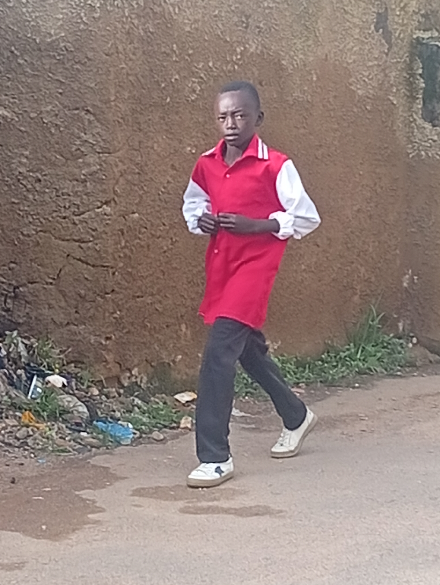 A young Black boy walks on a paved street in front of a rough, textured, brown wall. The boy wears a red collared shirt with white sleeves and collar accents, black pants, and white sneakers with blue accents. He is facing forward and appears to be walking towards the viewer's right. To the left of the boy, along the edge of the street and wall, is a small pile of debris and vegetation. The street surface is a mix of pavement and dirt, with some puddles of water. The scene suggests an outdoor urban environment in Jos, Nigeria.