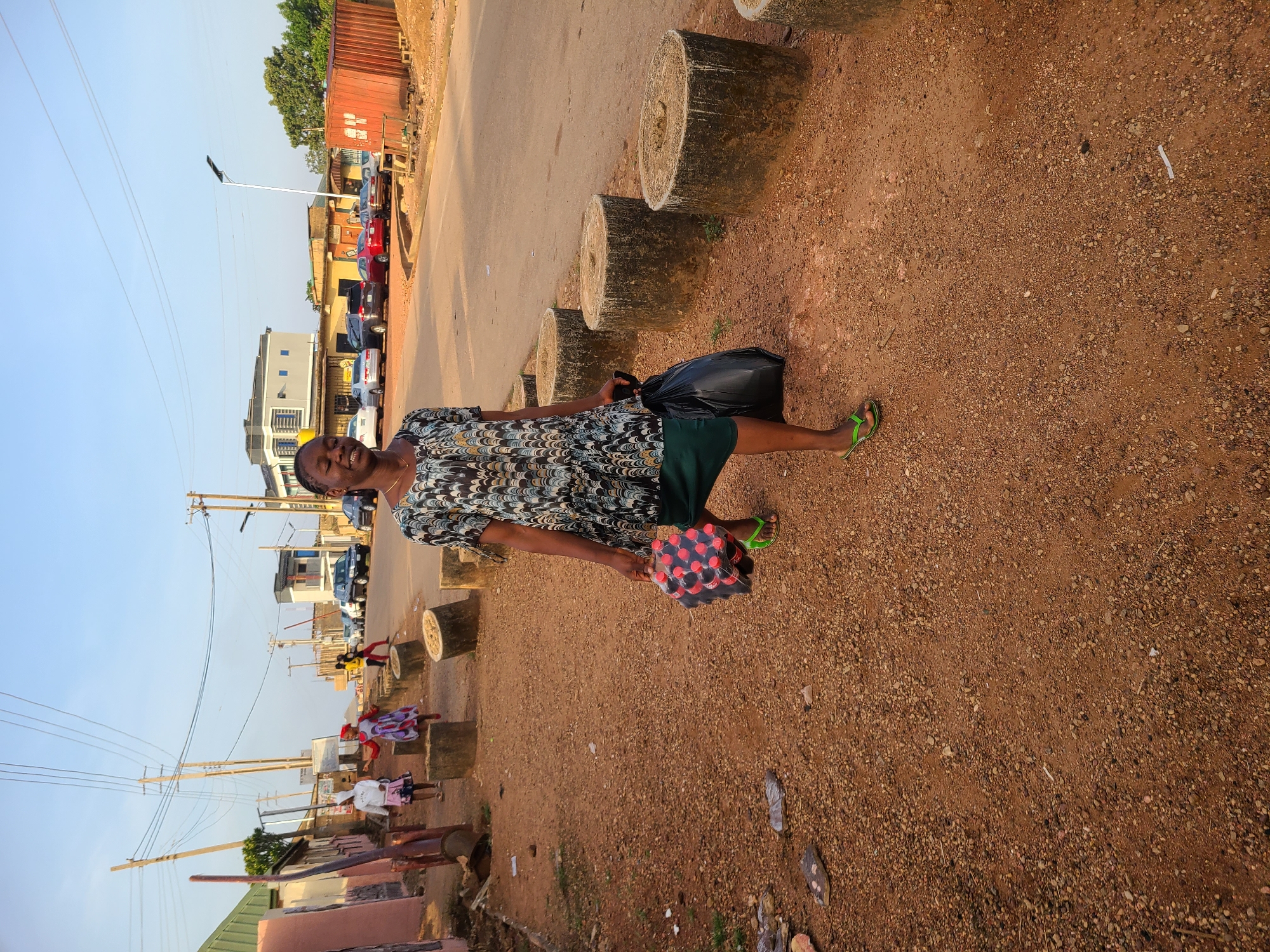 A person stands on a brown, rocky ground next to a street in Jos, Nigeria. They are carrying a black plastic bag and a package of red and black items. The person wears a patterned dress, green shorts, and green sandals. Beside them is a series of cylindrical concrete blocks separating the ground from the street. In the background are parked cars, buildings, and utility poles with wires. Other people are seen walking in the distance. A red shipping container is visible near the top of the image. The sky is clear and blue.