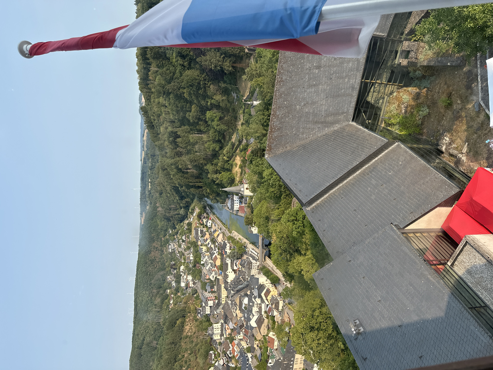 The image shows a panoramic view of Vianden, Luxembourg, taken from a high vantage point, likely the Vianden Castle. In the foreground, a flag of Luxembourg is visible, unfurled and waving. The flag has three horizontal bands: red, white, and light blue.
The middle ground displays a dense collection of buildings with red-tiled roofs, characteristic of the town of Vianden, nestled along the banks of the Our River. A bridge spans the river, connecting different parts of the town. A church spire with a white facade and dark roof is prominent among the buildings. The river winds its way through the valley, reflecting the surrounding greenery.
The background consists of rolling, tree-covered hills, typical of the Luxembourgish landscape. Wind turbines are visible on the crests of some hills, indicating a renewable energy presence. The sky is clear and blue, suggesting a sunny day. The architecture of the town, with its tightly packed houses and varied rooflines, speaks to its historical character. The overall impression is a scenic and tranquil European town situated within a lush natural environment.