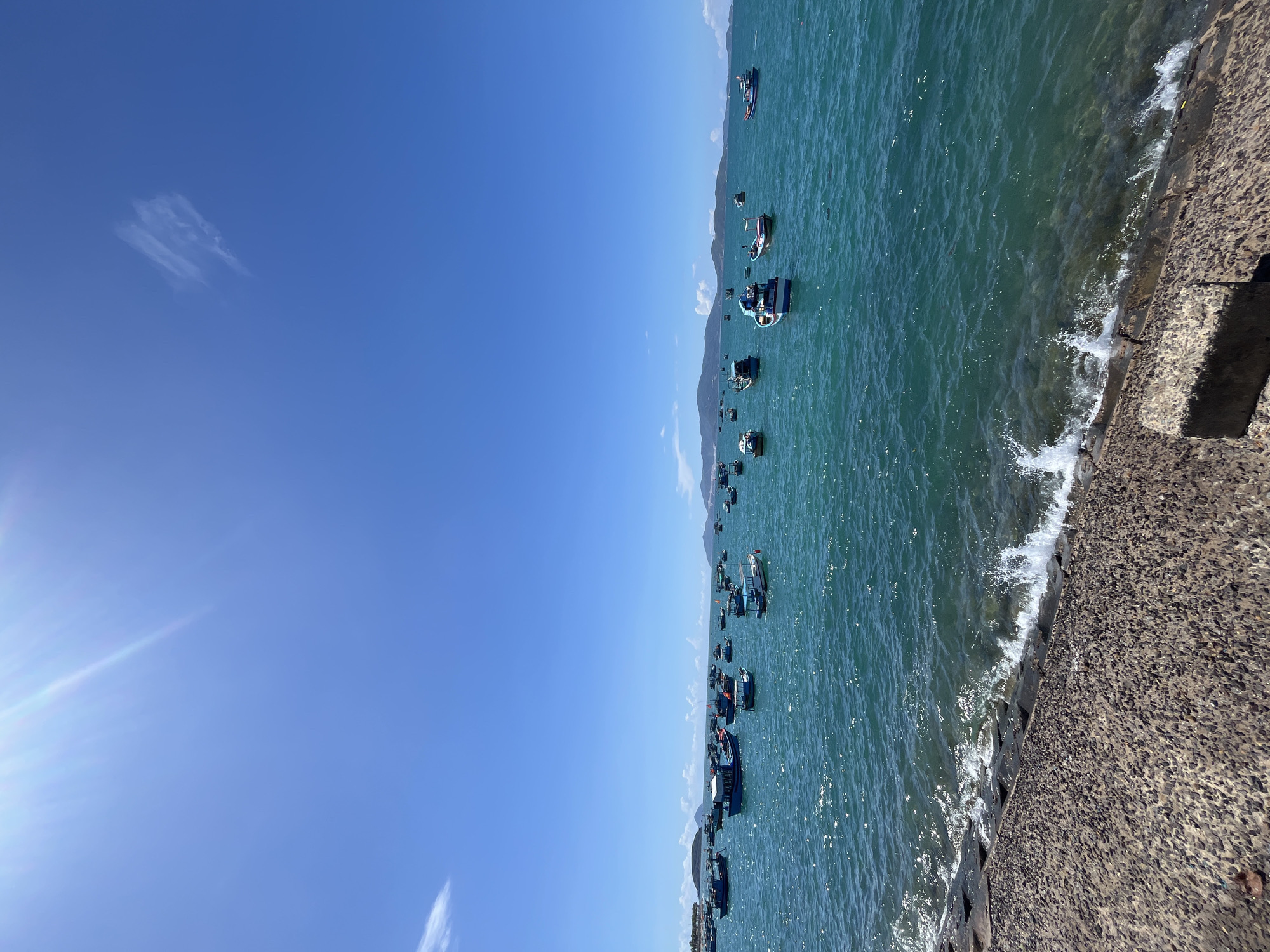The scene depicts a coastal view in Bac Nha Trang Ward, Vietnam. A bright blue sky with a few wispy clouds dominates the left side of the frame. The right side showcases the turquoise water of the sea dotted with numerous fishing boats. A landmass, potentially a mountain range, is visible in the distance. The foreground on the right consists of a concrete embankment where waves are crashing against the edge.