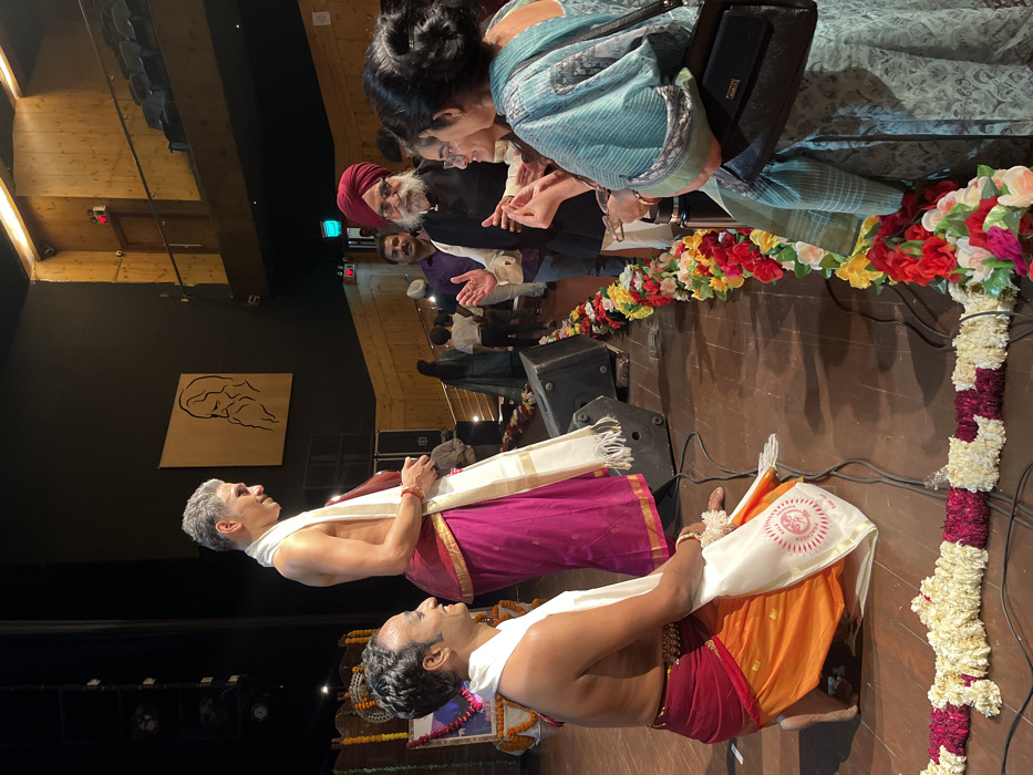 The image shows a group of people standing on a stage in a performance hall. The hall has a wooden ceiling and a black wall behind the stage, where a portrait of a bearded man is displayed. The scene is taking place in Chandigarh, India, at the coordinates 30.732 latitude, 76.789 longitude. Three men are on stage, one of whom is wearing a long pink and white dress and is kneeling on the stage floor. Another man, wearing a white kurta, is kneeling opposite him. An older man with a grey beard and white turban is standing behind the kneeling men, looking at the man in the pink dress. A woman wearing a teal dress and a black purse is standing to the right of the older man, looking at the man in the pink dress. A woman in a black dress is sitting on the right of the woman in teal. The stage is decorated with a long line of flowers laid down the middle. There are a couple of dark speakers in the background. The scene appears to be a performance or presentation, as the man in the pink dress is likely a dancer, and the older man appears to be a VIP guest or guest of honor. The women sitting and standing are likely attendees or audience members.