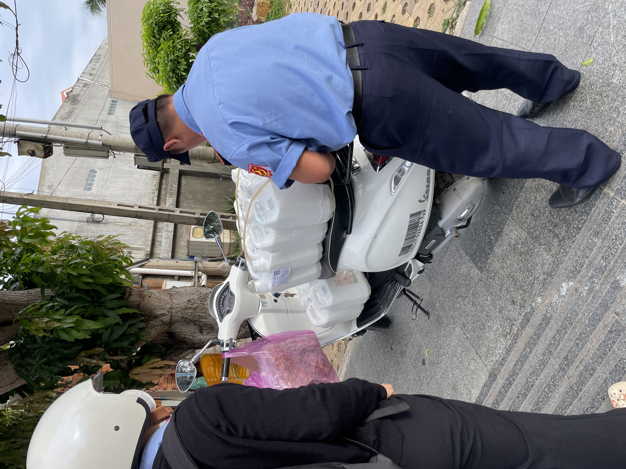 In Phu Thuy Ward, Vietnam, a person wearing a white helmet and black jacket stands near a white scooter. Another individual in a blue uniform, including a blue shirt, dark pants, and a dark cap, is attending to several white plastic containers loaded onto the scooter. A pink plastic bag containing what appears to be food items is also visible on the scooter. The background shows a building with a gray facade and a large tree. The ground is paved with stone.