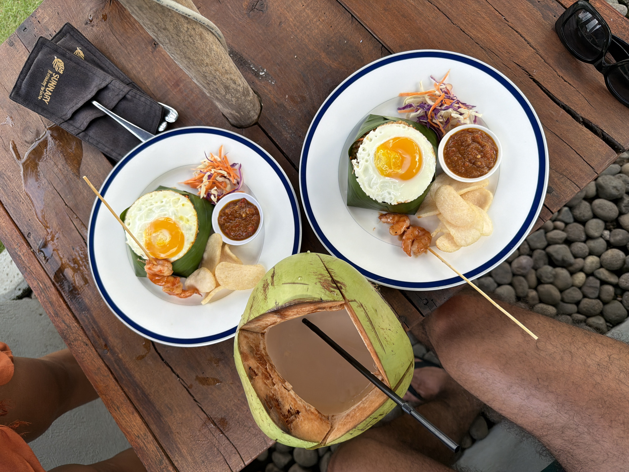 Two plates of food, each containing a fried egg, what appears to be rice wrapped in a leaf, a small bowl of sauce, a skewer of grilled shrimp, and some chips, are placed on a wooden table. Next to the plates is a halved green coconut with a black straw inserted. A black sunglasses case and silverware are on the table near one plate. The table is outdoors, and in the background, there are gray pebbles visible. Two people are at the table, one wearing an orange garment and the other dark shorts and flip-flops. The image was taken in Selemadeg Timur, Indonesia.