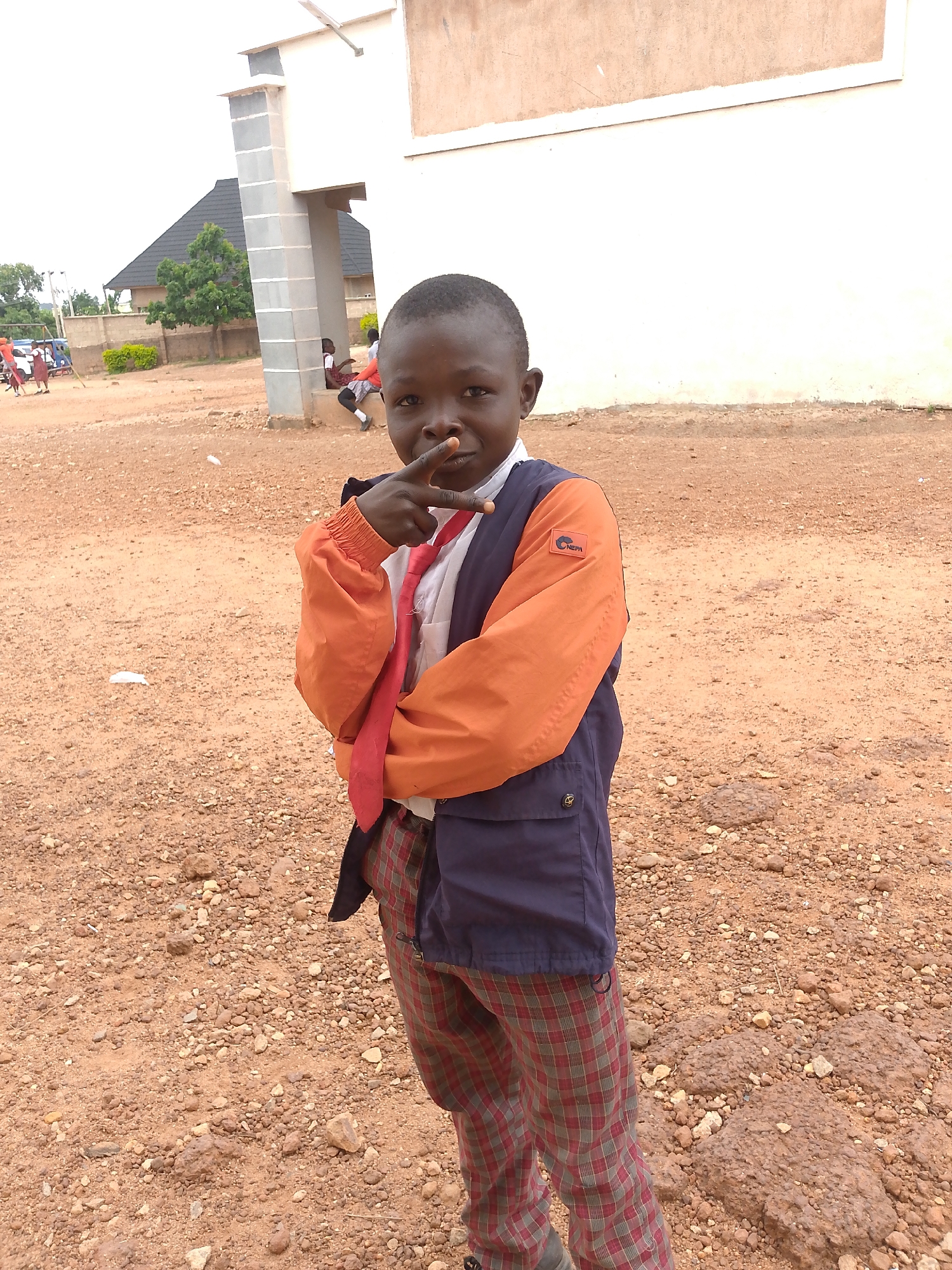 A young boy stands outdoors on a reddish-brown gravelly ground. He is wearing a dark blue vest over an orange jacket, a white shirt, a red tie, and plaid pants. His left hand is raised, with his index and middle fingers extended in a peace sign towards the camera. In the background, a light-colored building with a darker roof is visible, along with trees and other people in the distance, suggesting an urban or suburban setting. The overall scene appears to be in Bukuru, Nigeria, a town located in Plateau State.