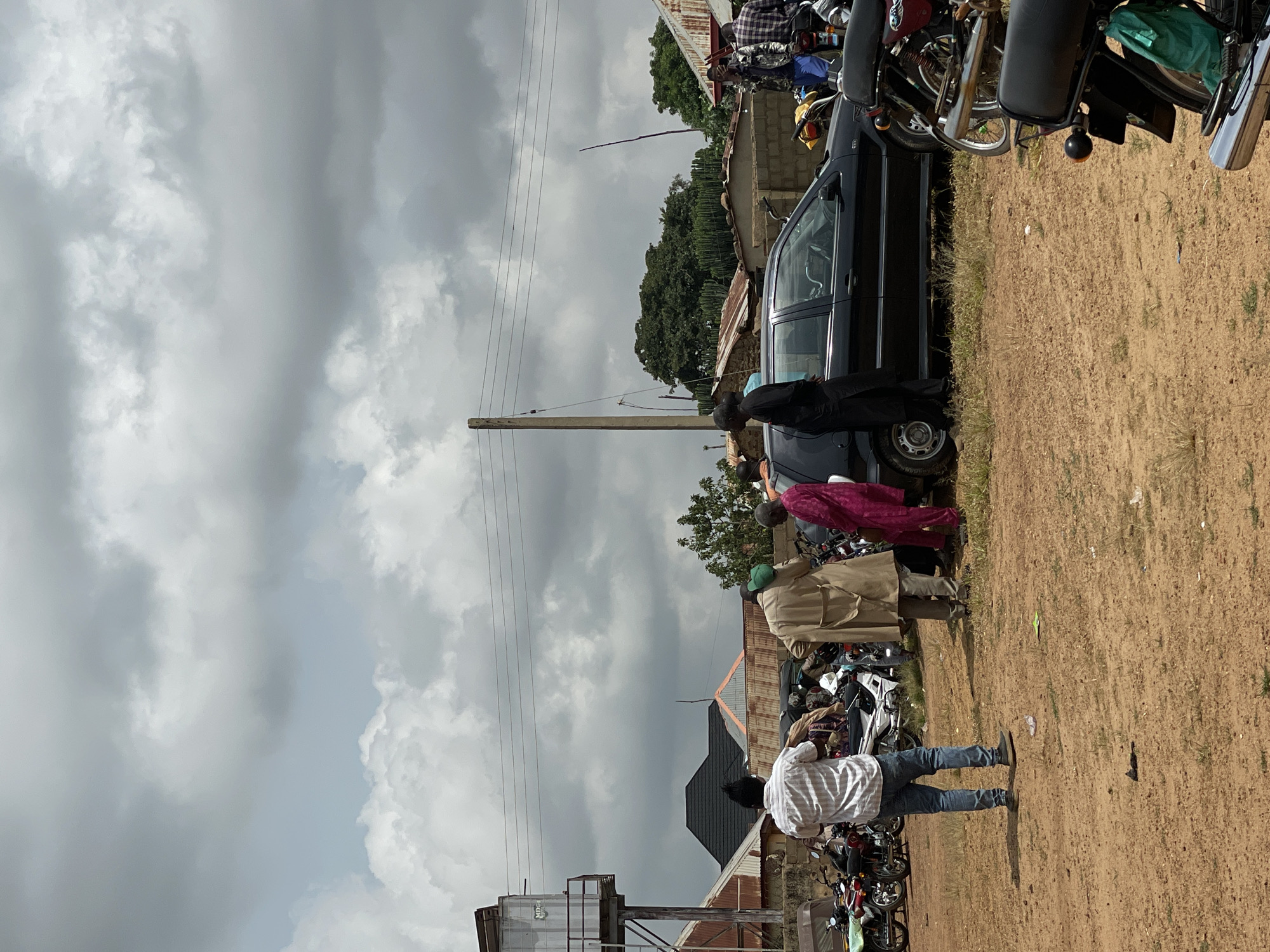 The image depicts an outdoor scene in Kassa, Nigeria, under a cloudy sky. Several people are gathered around a black sedan and a collection of motorcycles.
In the foreground, the ground is composed of reddish-brown dirt with sparse patches of dry grass. A man in a white striped shirt and blue jeans stands facing away from the camera, his back to a group of motorcycles. To his right, a man in a tan coat stands with his back to the camera, holding a green object in his left hand. A woman in a bright pink garment is to his right, facing the car. Another person in black attire is standing on the other side of the car.
The black sedan is parked in the middle ground, with its doors slightly ajar and its front wheels turned. A row of motorcycles is parked to the left of the sedan. Further to the left, a water tower structure is visible, with corrugated metal roofing on adjacent buildings.
In the background, more buildings with corrugated metal roofs are visible, interspersed with trees. A wooden utility pole with wires runs across the scene. The sky is filled with heavy, grey clouds, suggesting an overcast or potentially rainy day. The overall impression is one of a community gathering or a temporary stop in a semi-urban or rural area.