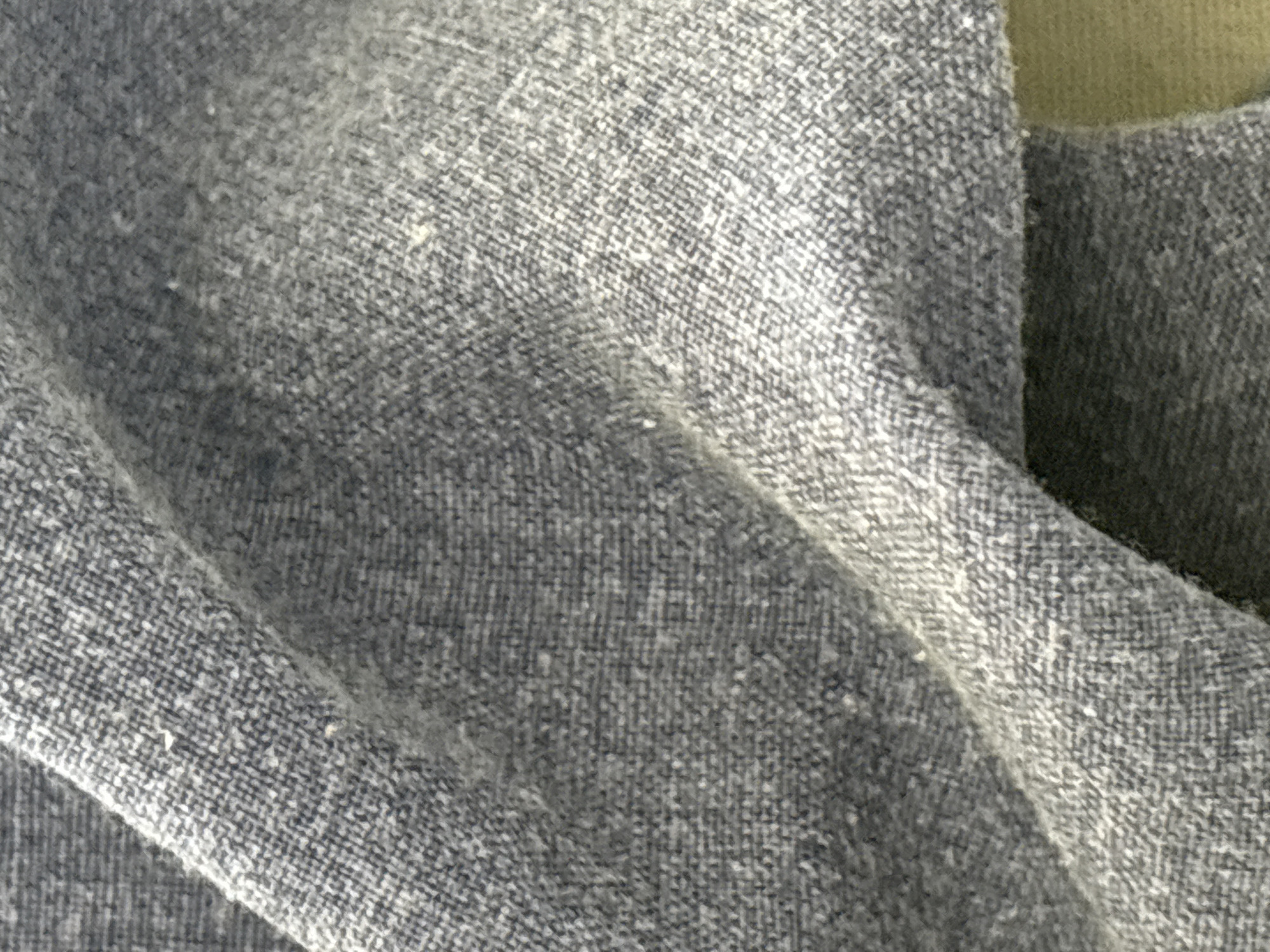 The image is a close-up, overhead shot of a wrinkled piece of textured, grey fabric. The fabric appears to be a woven material with visible threads creating a subtle pattern. The lighting creates shadows and highlights, emphasizing the folds and texture of the cloth. The background is out of focus and a darker shade of grey or black. There are no people, animals, or identifiable objects in the image. The location is unknown.