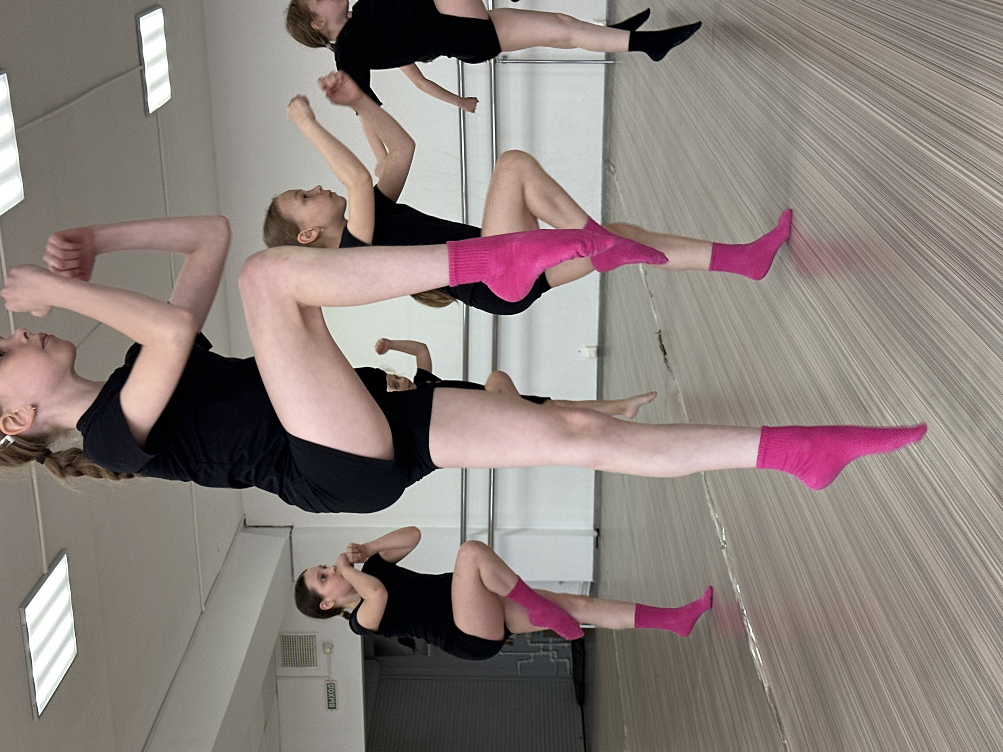 The image depicts a dance class in Votkinsk, Russia. Several young girls, dressed in black leotards and shorts, are practicing dance moves. Some girls are wearing pink socks, while at least one girl wears black socks. The room appears to have mirrors on the walls, reflecting the dancers' movements. The lighting is provided by rectangular ceiling lights. An exit sign, written in Russian "ВЫХОД" is visible on the left side of the frame. The floor is a light-colored, horizontally-lined material.