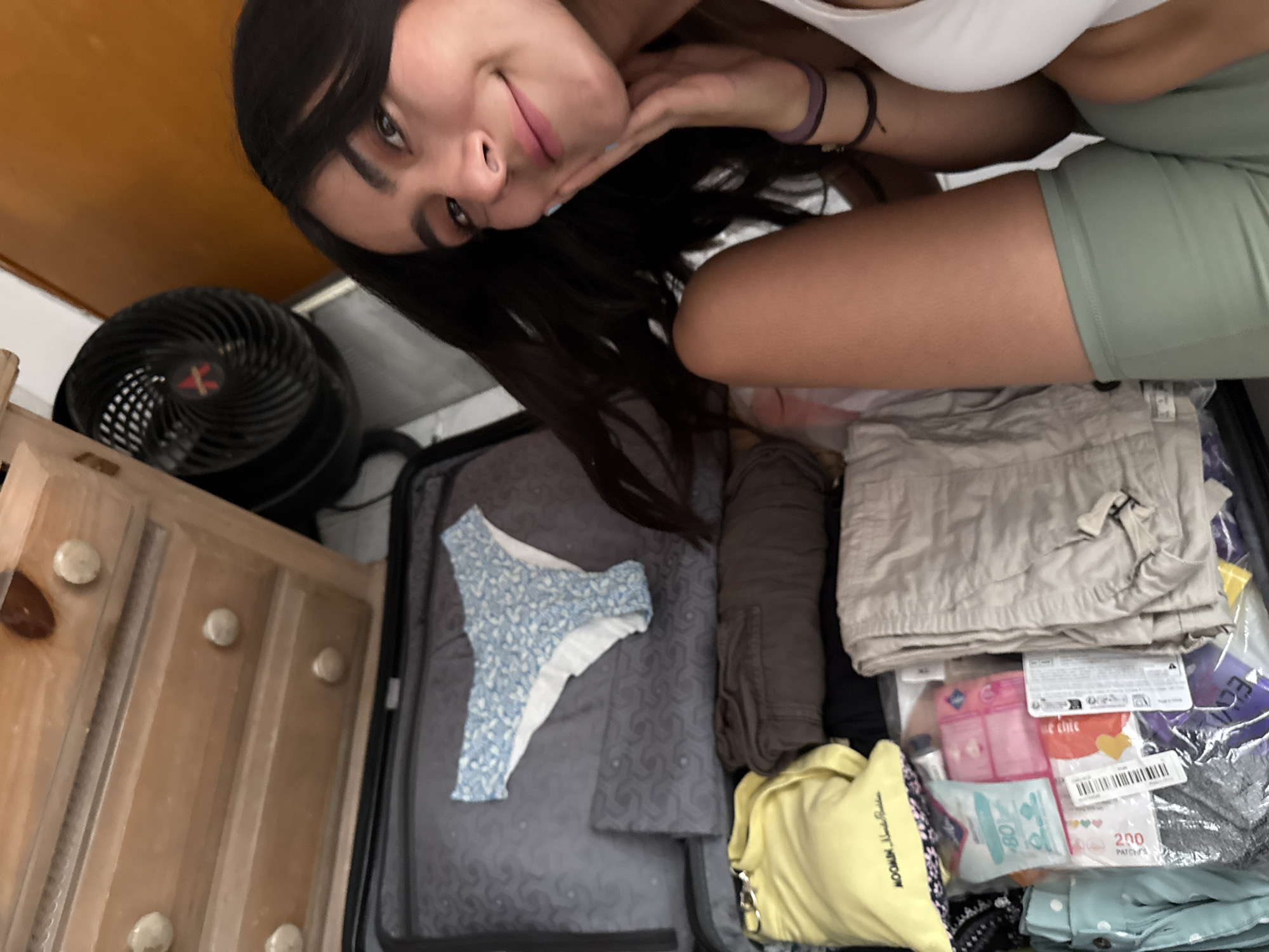 A woman with long dark hair is posing, partially visible, above a suitcase packed with clothing. The suitcase contains: a blue and white patterned swimsuit bottom, folded khaki pants, rolled brown clothing (likely pants), a yellow shirt, and a clear plastic bag containing what appears to be travel-sized toiletries and first aid items. A black fan is visible to the left of the suitcase, adjacent to a wooden dresser. The location is presumably indoors, possibly in Puebla, Mexico.