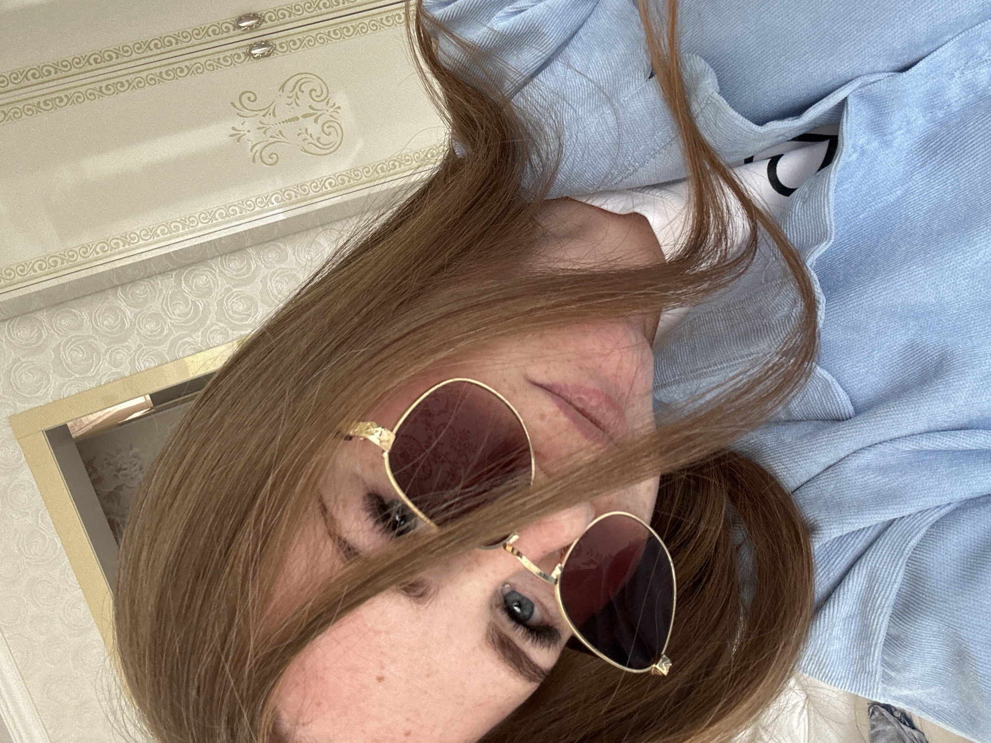 A person with light brown hair and sunglasses is lying on a surface, possibly a bed or sofa, wearing a light blue garment, perhaps a shirt or jacket. A decorated box with gold accents is visible in the upper left portion of the image. It is impossible to provide any location context without additional information.