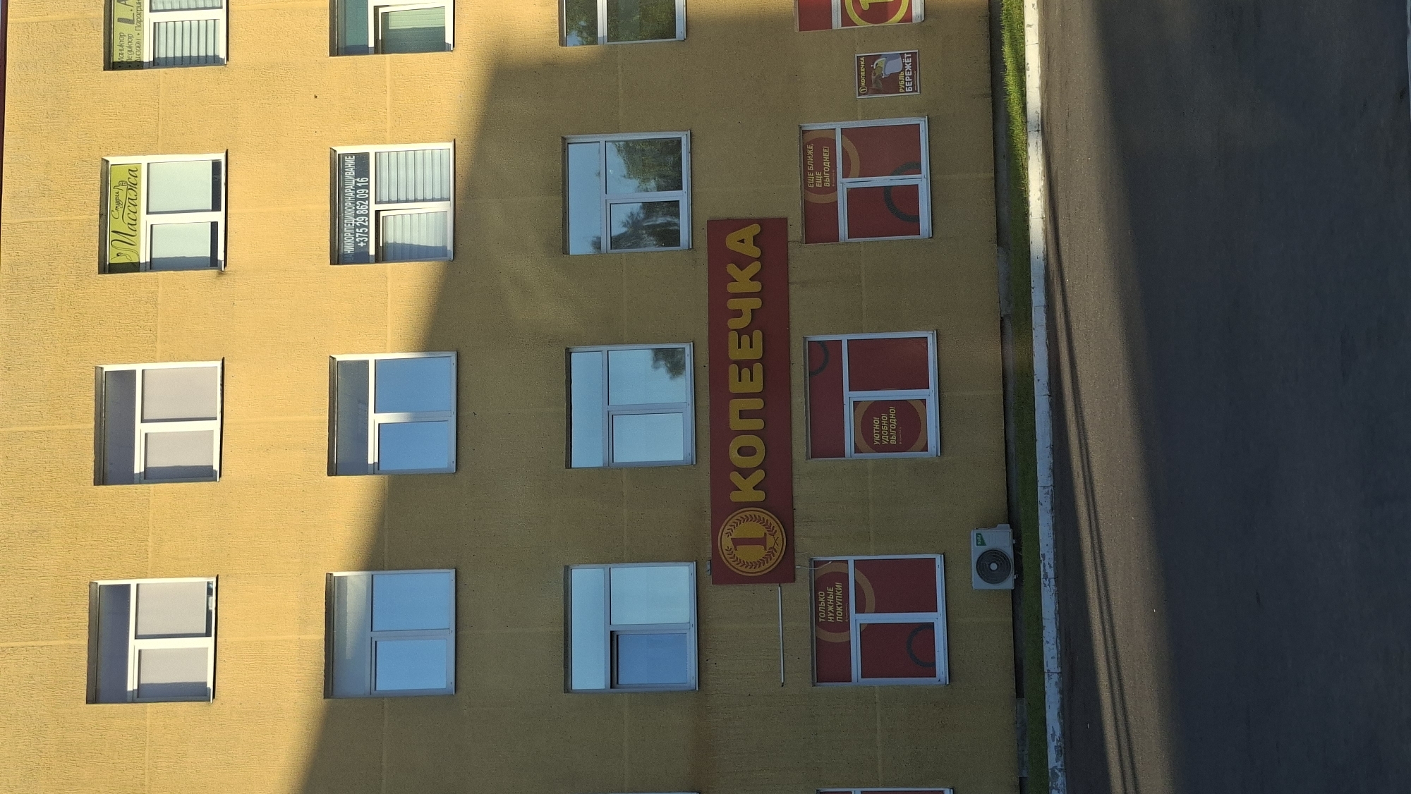 The image shows the facade of a yellow building with multiple windows. One prominent sign on the building reads "КОПЕЕЧКА" in large, yellow, capital letters, with a circular logo featuring a laurel wreath and a coin with the number "1" on it. Below this main sign, there are several windows with red and white advertisements. One advert shows a coin and text "РУБЛЬ БЕРЕЖЕТ" (Ruble saves). Another advert has text "УДОБНО! УДОБНО! ВЫГОДНО!" (Convenient! Convenient! Profitable!). Another advert says "ТОЛЬКО НУЖНЫЕ ПОКУПКИ!" (Only necessary purchases!). There are also smaller signs on windows higher up the building, including one for "Массаж" (Massage) and another for "МАНИКЮР ПЕДИКЮР НАРАЩИВАНИЕ" (Manicure Pedicure Extension) with a phone number. An outdoor air conditioning unit is visible near the bottom of the building. The building appears to be located in the city of Smalyavichy, Belarus, as suggested by context provided with the image. The sun is casting a strong shadow across the building and the pavement in front of it.