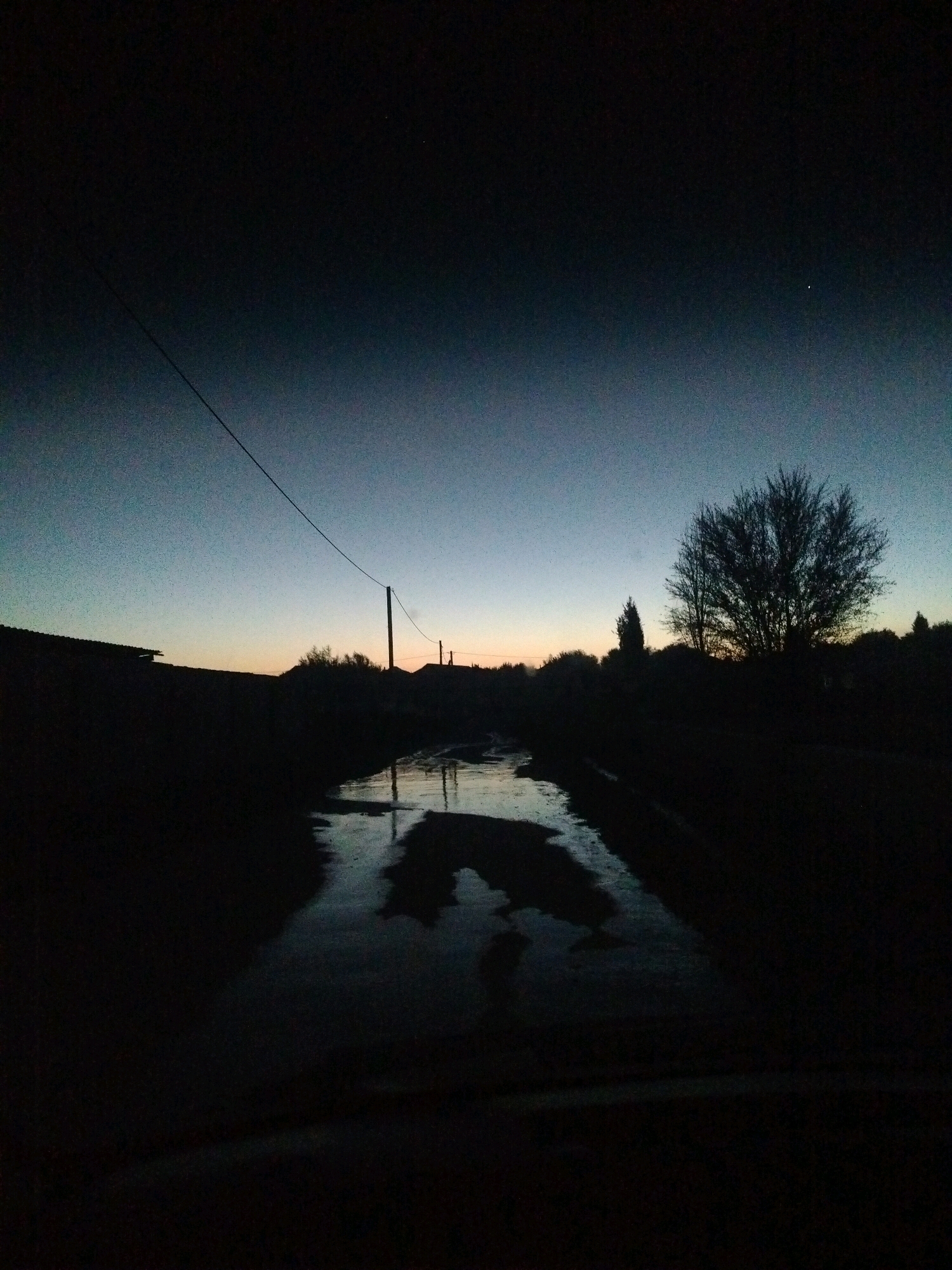 The image depicts a dark, rural scene, likely at dusk or dawn, with a clear gradient in the sky from deep indigo at the top to a lighter, almost pale blue towards the horizon. A single bright star or planet is visible in the upper right quadrant of the sky. A power line stretches diagonally from the upper left to a utility pole situated roughly in the center of the frame, to the right of a low-lying structure on the left.
In the foreground, a body of water, possibly a flooded road or ditch, reflects the dim sky and surrounding dark silhouettes. The reflections are somewhat distorted, suggesting ripples or uneven surfaces on the water. On the left side of the frame, the dark silhouette of a building with a slanted roof is visible. To the right of the water, silhouetted trees and bushes line the landscape, with a larger, bare tree standing prominently towards the right side of the image. Distant, smaller silhouettes of structures or trees are also present along the horizon. The overall impression is one of quiet solitude and a transition from day to night in a possibly undeveloped or semi-rural area. The specific location in the city of Көлкент, Kazakhstan, is not visually evident from the provided image alone.