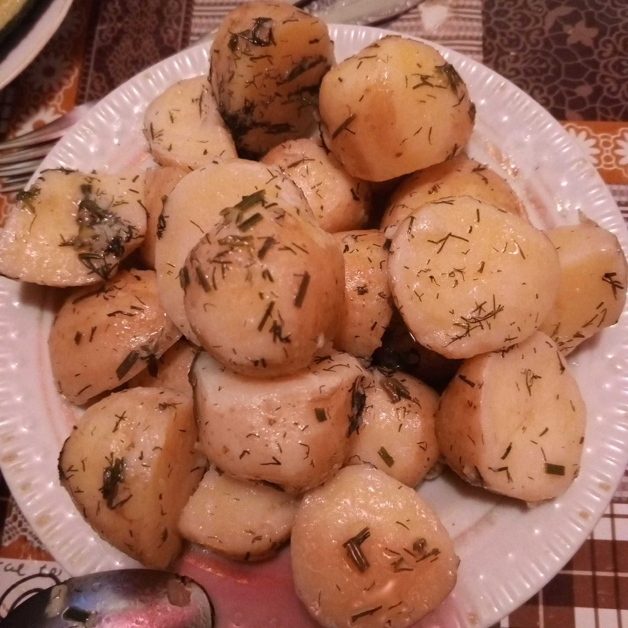 A close-up, overhead shot shows a white, ruffled plate filled with boiled potatoes. The potatoes are small and round, some appearing to be halved or quartered. They are seasoned with chopped fresh dill, visible as dark green flecks scattered across their golden-yellow surfaces. The potatoes are glistening, indicating they may be served with butter or oil. The plate rests on a patterned tablecloth in shades of brown and orange, with a checkered pattern visible on the right side. A fork is partially visible on the left side of the frame, suggesting the dish is ready to be eaten. The lighting is warm and the overall scene evokes a sense of home-cooked comfort. No specific location or city is discernible from the image.