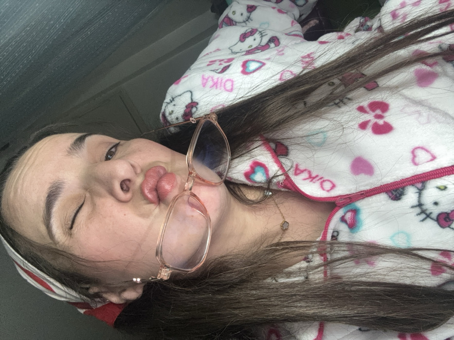 A young woman in Hello Kitty pajamas winks, her lips puckered. She's wearing rose-colored glasses perched on her nose. A matching headband is visible. Her hair is long and brown. A clover necklace is around her neck. The pajamas have pink accents including hearts and the "Dika" brand name written on them. The scene is casually lit, suggesting an indoor setting.