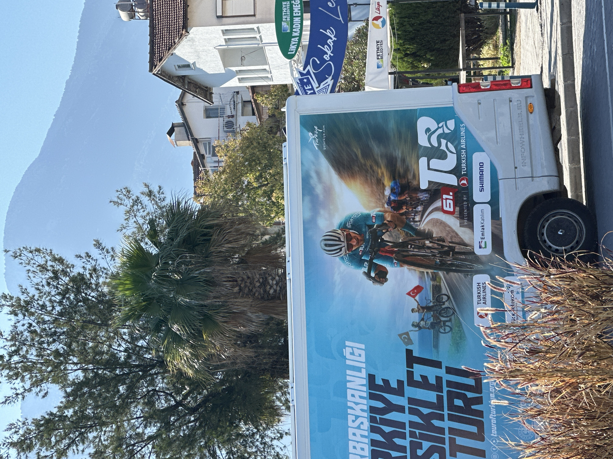 An outdoor daytime scene in Fethiye, Turkey, features a white promotional truck or van parked alongside a street or sidewalk. The vehicle's side is covered by a large graphic advertising the "TÜRKİYE BİSİKLET TURU" (Tour of Turkey Cycling), marking its 61st edition. The graphic prominently displays a dynamic image of a cyclist, clad in a teal and blue jersey and helmet, riding a bicycle. Below this, smaller illustrations show individuals on bikes holding Turkish flags.
Numerous sponsor logos are visible on the graphic, including "TURKISH AIRLINES," "SHIMANO," "Emlak Katılım," "Türk Telekom," and "SPORTOTO." Other text on the advertisement reads "Türkiye goturkiye.com" and "tourofturkiye.org." On the rear of the actual vehicle, "INFOWHEELS.EU" is embossed.
In the background, under a clear, bright blue sky, residential buildings with white facades and terracotta roofs line the street, some featuring balconies. A large mountain range is visible in the distance behind these structures. The foreground and sides are populated by lush green foliage, including palm trees and other dense trees, along with some drier, brush-like plants. Several vertical banners are also present, displaying text such as "FETHİYE BELEDİYESİ" (Fethiye Municipality), "LİKYA KADIN EMEĞİ," and partially visible "Sokak Le..." and "Likya," providing local context. The time of day appears to be bright and sunny with clear weather.