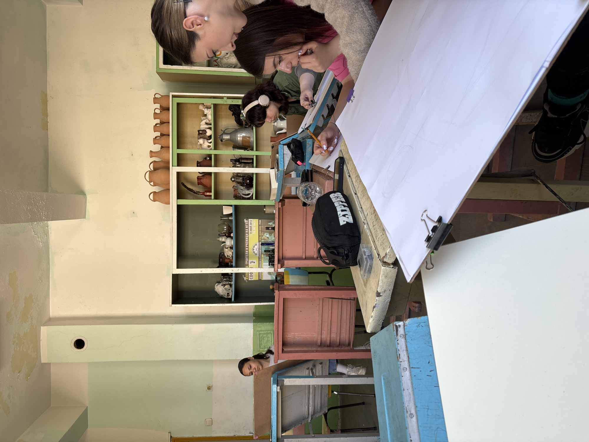 This media file captures an indoor scene, likely an art classroom or studio, in Troyan, Bulgaria. The primary subjects are three young women, concentrated on drawing or sketching. Two are in the immediate foreground, leaning over large white sheets of paper on tables, holding pencils. One has headphones on. Their expressions suggest focus and engagement in their creative task. A third woman is visible further back, peering over another table, also seemingly involved in the activity.
The setting features light green or beige walls, which show signs of wear and tear with some peeling paint. In the midground, several wooden shelves are filled with art-related objects. These include rows of small terracotta pots, various ceramic figurines such as sheep or lambs, and metal vessels like pitchers, arranged as potential still-life subjects. The furniture consists of rustic-looking wooden tables and chairs, some painted in shades of red-brown and blue. A black backpack and cell phones are also visible on the tables.
On one of the shelves, a poster is displayed with partially visible Cyrillic text. The legible parts read "УЧИТЕЛИ И УЧЕНИЦИ НА ПЕ" (Teachers and Students of Pe...) and "15 МАР 2012", indicating an event date of March 15, 2012. The text "СОО" is also visible. The lighting suggests it is daytime, creating a functional atmosphere for an art lesson or workshop.