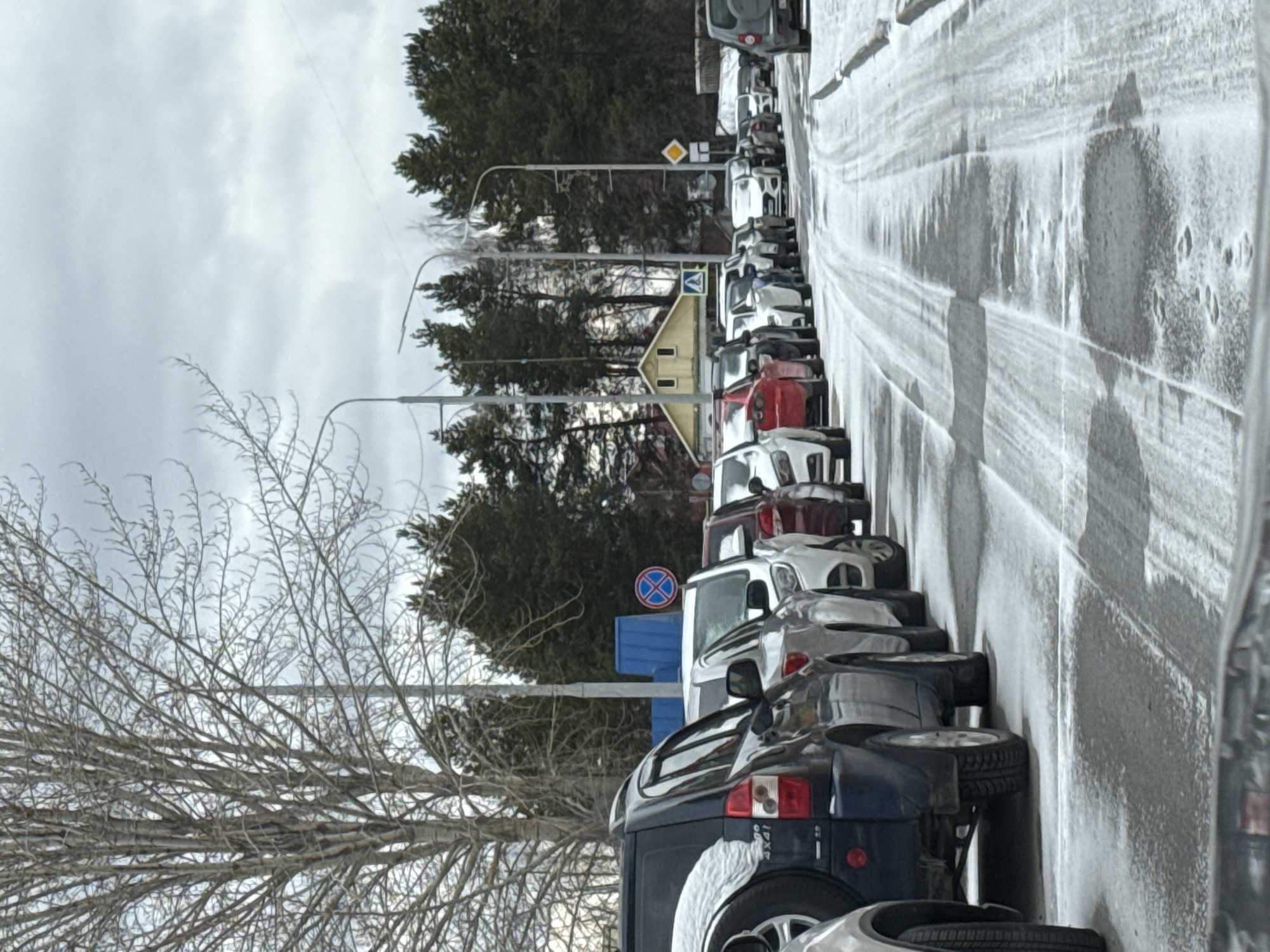 The image shows a line of cars parked on the side of a snowy road in Beloyarsky, Russia. The cars are parked close together, facing in the same direction. There are trees and buildings in the background. The sky is overcast. A bare tree is visible in the foreground. Several road signs are present along the road. The road is icy.
