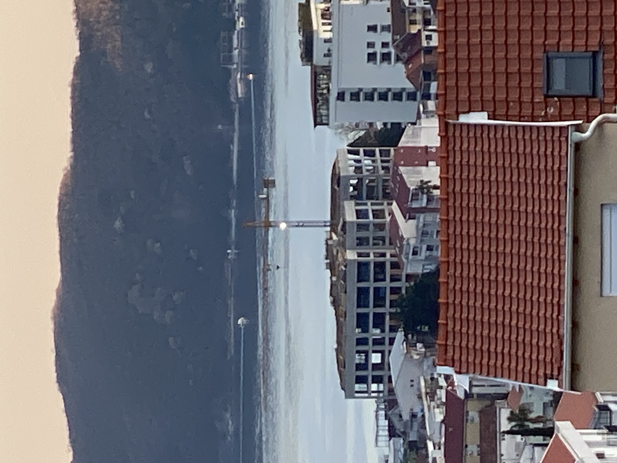 The image depicts a cityscape of Budva, Montenegro, with a body of water visible in the background. A coastline, possibly a bay, separates land with denser vegetation from buildings and infrastructure. Predominantly white buildings with visible windows and architectural details rise amongst the red-tiled roofs typical of the region. A construction crane is also visible near the water. The buildings exhibit varied heights and configurations, indicating a mixed urban development. The picture is taken from an elevated perspective, providing a wide view of the city and its surroundings.