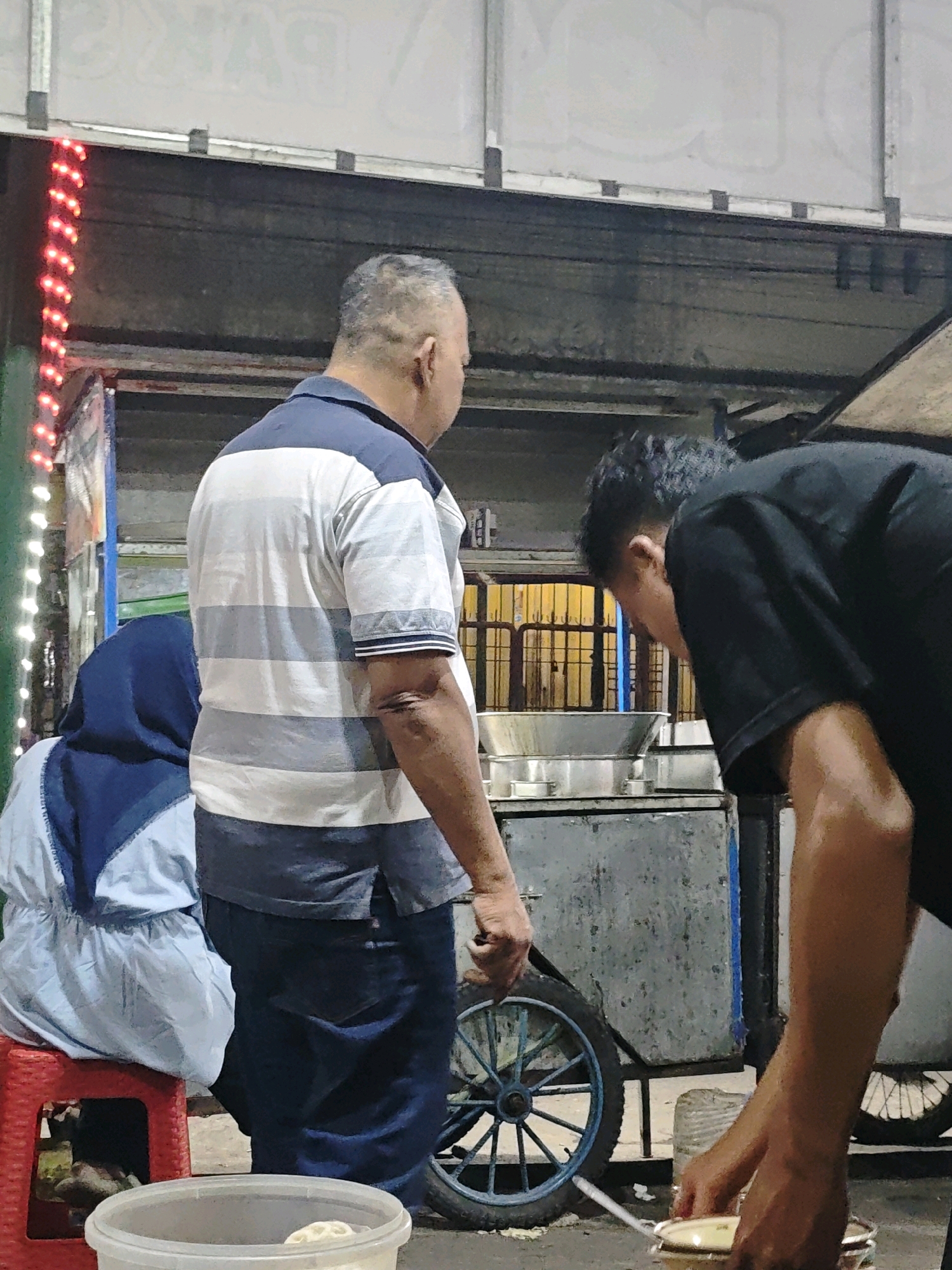 In Ngasem, Indonesia, at night, three people are near a food cart. A man in a striped polo shirt and dark pants stands facing away from the camera, looking towards the cart. To his left, a person wearing a blue hijab and light blue clothing sits on a red stool, also facing the cart. In the foreground, another man in a black t-shirt is bending over, reaching towards a white bowl. The food cart has a large metal basin on top, and a blue spoked wheel is visible on its side. Red lights are strung along a structure to the left of the scene. The overall environment appears to be an outdoor market or street food stall.