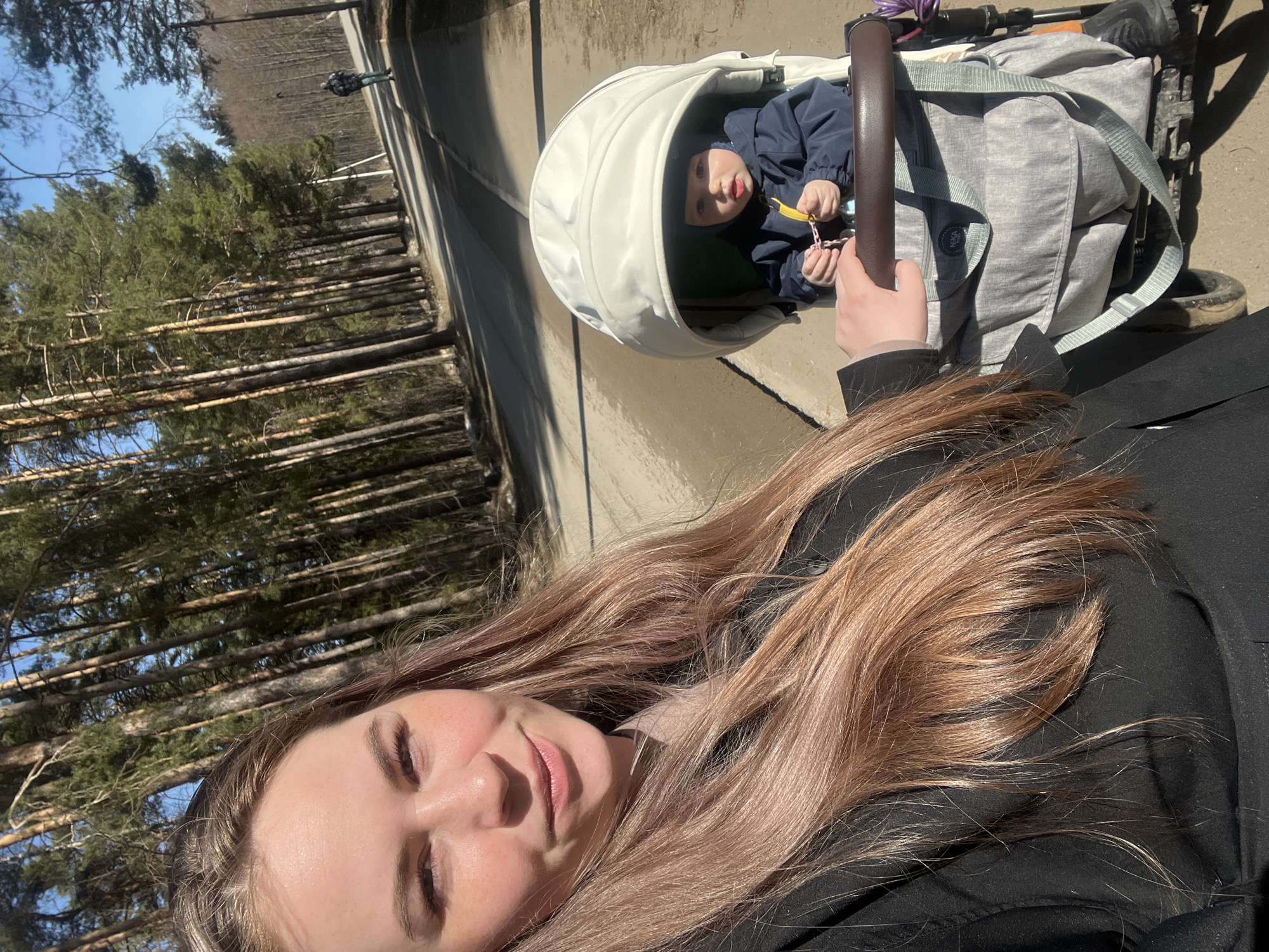 A selfie taken by a woman with long brown hair, wearing a black jacket, shows her standing next to a baby in a white stroller in Chaikovsky, Russia. The baby wears a blue jacket and holds a yellow object in their hands. The stroller is on a concrete surface, beside a dense forest of tall, thin trees on a sunny day. A road and a grassy area are visible in the background. The stroller has a gray bag attached to it.