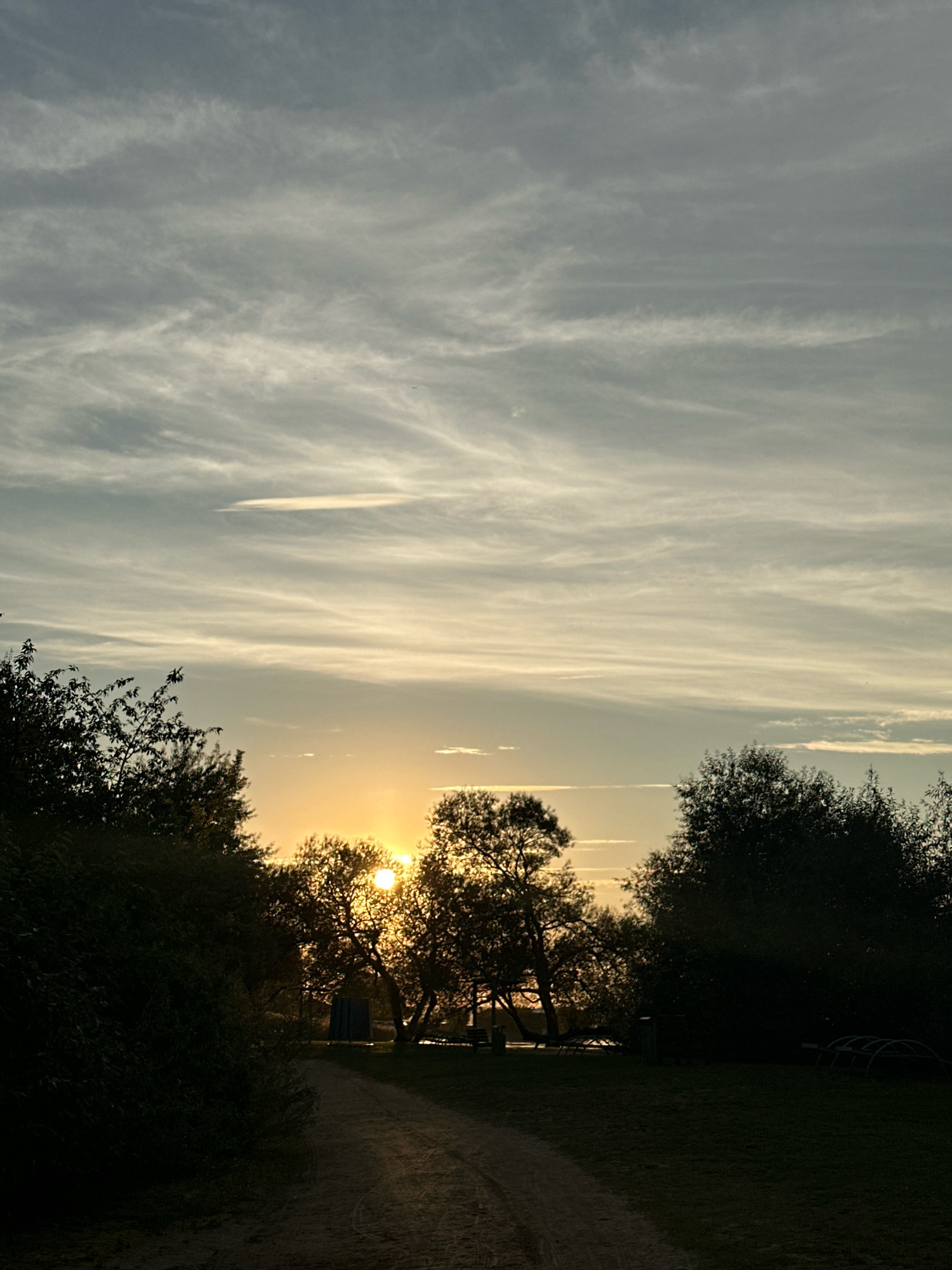 A dirt path curves into a park-like area at dusk. Silhouetted trees line the sides of the path, with the setting sun visible between them, casting a warm glow. The sky is filled with wispy clouds, indicating a transition from day to night. In the background, there's a hint of a building or structure, and some playground equipment, suggesting a public space. The overall scene is peaceful and natural.