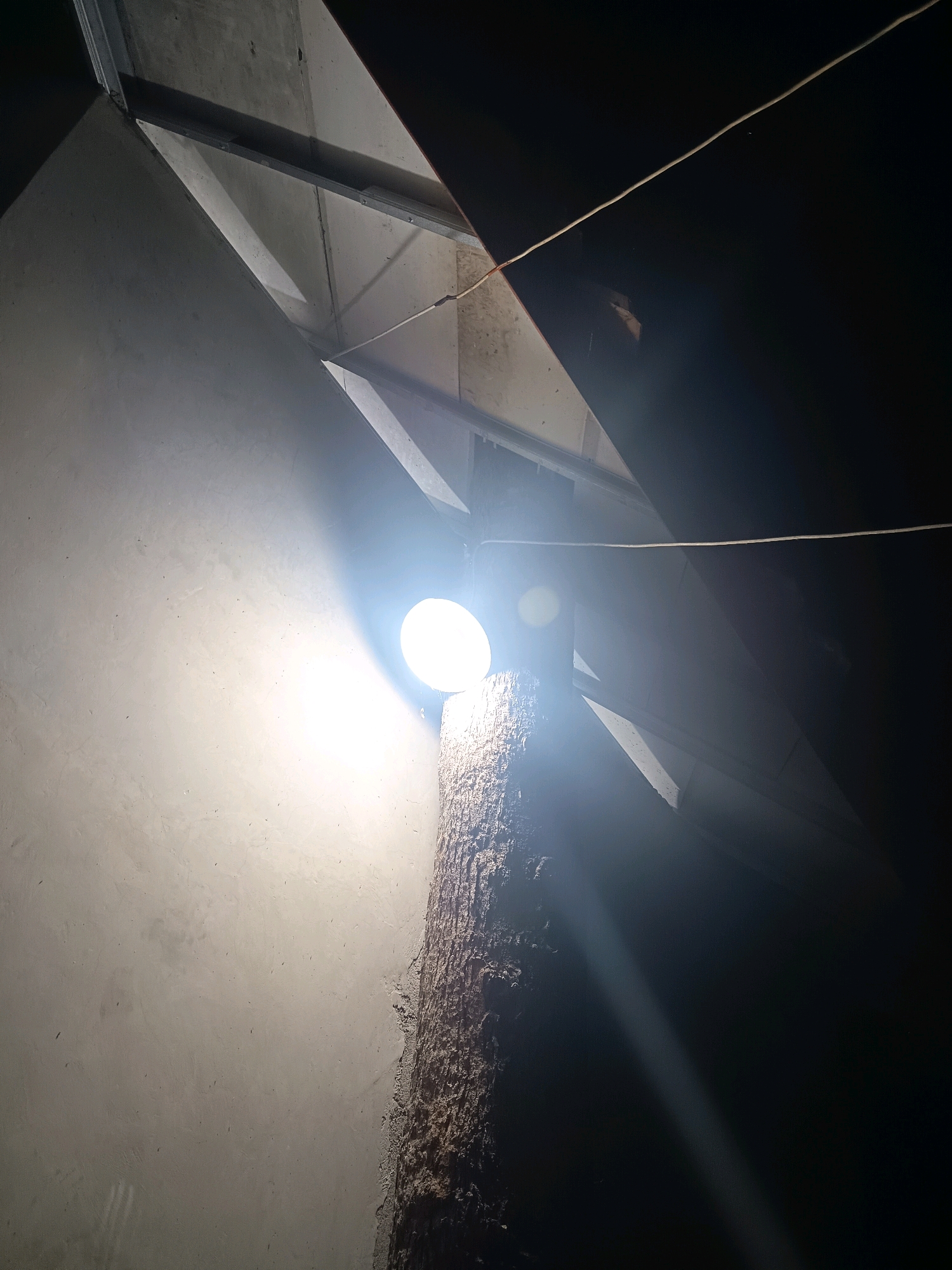 A bright, circular light bulb is illuminated and attached to a rough, vertical beam, possibly a piece of wood. The beam is positioned in front of a textured, light gray wall. Above the beam and wall, a ceiling structure is visible, composed of light-colored panels and dark, angular supports. A thin, light-colored wire stretches diagonally across the upper portion of the image, disappearing into darkness on the right. The lighting creates stark contrasts, with the area around the bulb brightly lit and the background on the right side of the image completely dark. There are no people or animals visible in the scene. This image appears to be taken indoors, possibly in a construction or unfinished space, and does not contain any specific elements that would definitively place it in Sumedang, Indonesia.