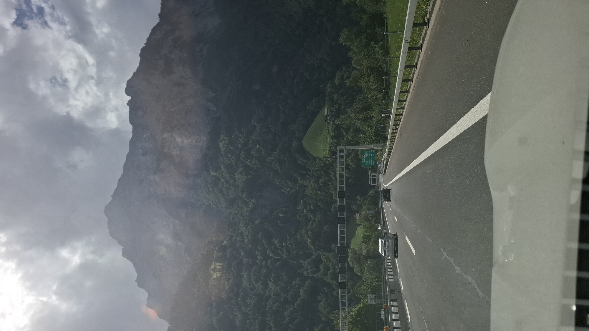 This image captures a view from a vehicle traveling on a highway in Seedorf (UR), Switzerland. A massive, rugged mountain looms in the background, its rocky face partially obscured by atmospheric haze and dramatic clouds. The mountain is densely covered with lush green forest at its lower slopes, with a small, bright green field and a few buildings visible higher up.

The highway itself is asphalt with clear white lane markings. Several cars are visible on the road, including a silver car in the left lane and a dark car in the right lane. Overhead gantries with electronic displays span the highway, and road signs with destination information are present. The foreground shows the dashboard or hood of the vehicle from which the photo is taken. The overall environment suggests a scenic drive through a mountainous region.