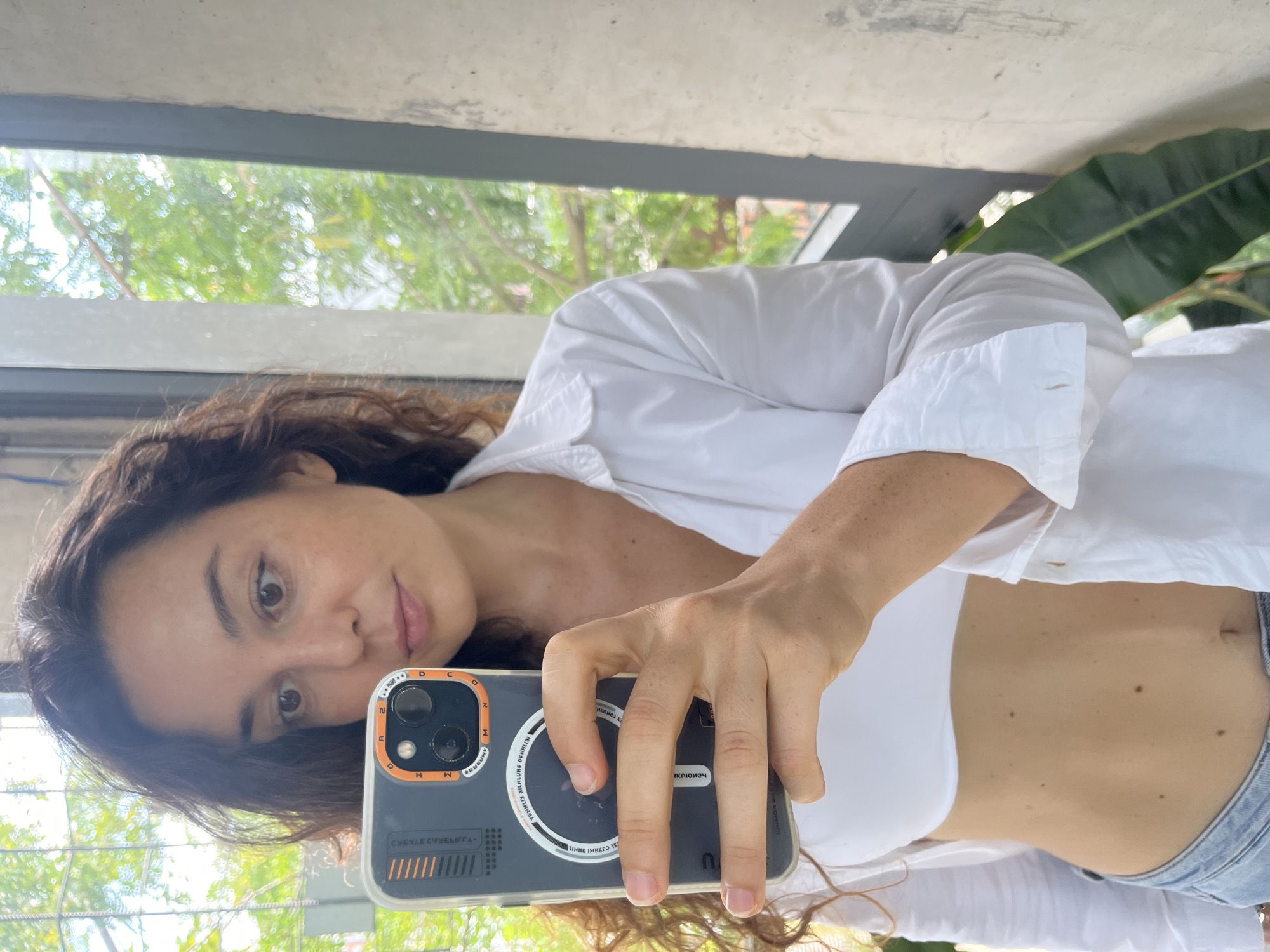 A woman with dark, curly hair is taking a selfie in what appears to be an apartment with a large window overlooking lush greenery. She is wearing a white crop top and an open white collared shirt, with light-wash jeans visible at the bottom of the frame. Her left hand holds a smartphone, the camera facing her. The phone has a dark case with orange accents. The background shows a concrete wall to the right and a blurred view of trees and foliage through a window to the left. The lighting suggests it is daytime. The location is identified as Phu Thuy Ward, Vietnam.