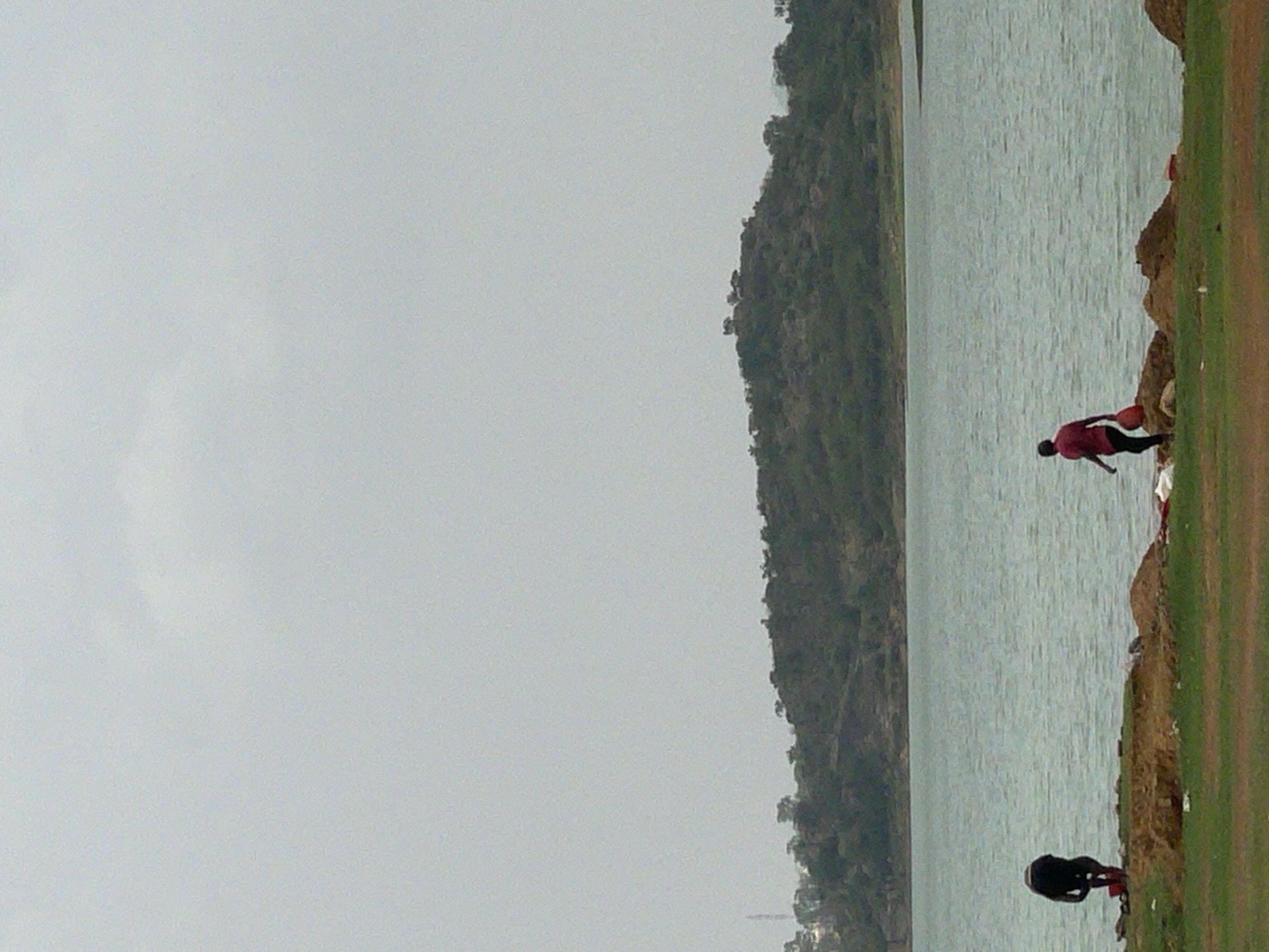 The image shows a body of water with a grassy bank on the right side of the frame. Two people are visible near the water's edge. One is wearing a red shirt and dark pants and is bent over, possibly interacting with something near the water. The other is wearing dark clothing and is also near the water. In the background, across the body of water, is a densely wooded hillside. The sky is overcast and grey. Given the provided information, this scene could be located in Nabatum, Nigeria.