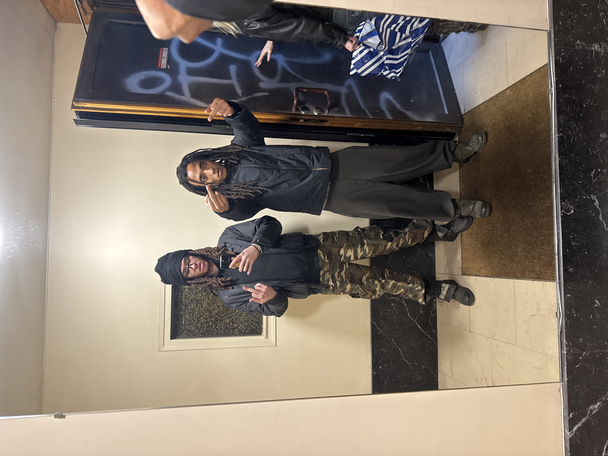 Two individuals are reflected in a mirror within an interior space. The person on the upper portion of the mirror wears a black jacket and dark-colored trousers, gesturing with their hands. The individual below is clad in a black jacket and camouflage pants, also gesturing. The surrounding environment includes a door with graffiti, light-colored walls, and a floor with a mix of white tiles, brown carpeting, and a black marble pattern. The image does not provide information indicating a location within Paris, France.
