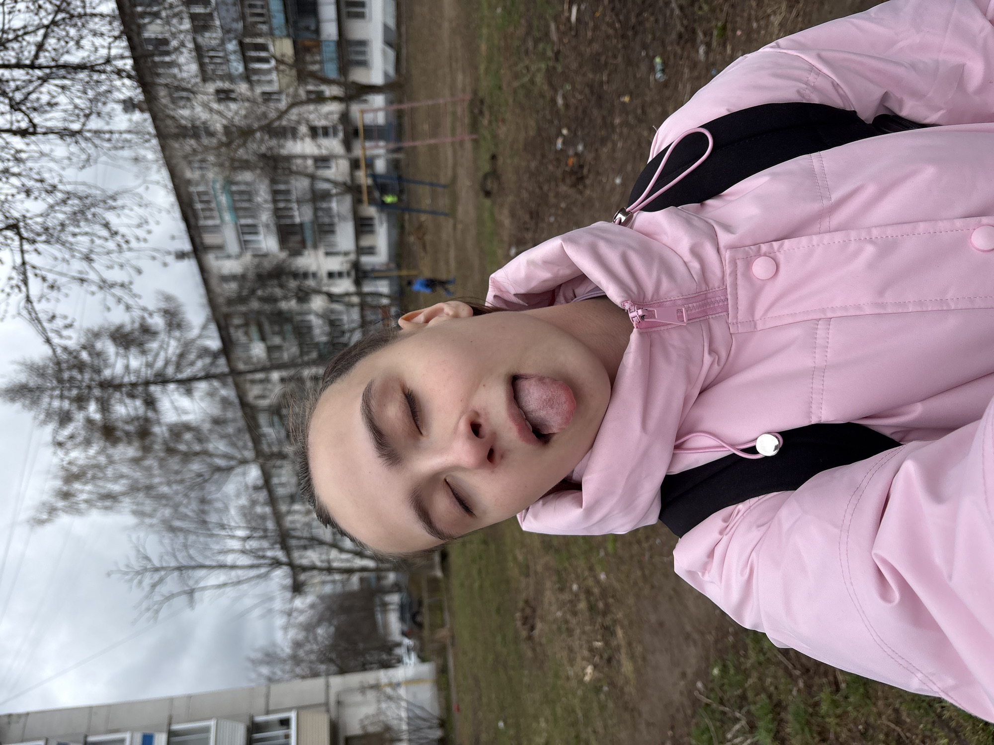 The image captures a young person, likely female, in a playful pose outdoors. Their eyes are closed, and their tongue is sticking out, conveying a lighthearted and humorous expression. They are wearing a bright pink puffer jacket with a visible black strap underneath, possibly from a backpack. The jacket features a prominent pink zipper, snap buttons, and a drawstring with a silver toggle.
The background depicts an urban or suburban residential area, consistent with Chaikovsky, Russia. Several multi-story apartment buildings are visible, some with multiple windows and subtle blue-tinted sections. The foreground and mid-ground show bare trees, characteristic of a colder season like late autumn, winter, or early spring. The ground is unkempt, covered in dry grass and dirt. Faintly visible in the distance are elements of a playground structure, including what appears to be horizontal bars. The sky is overcast, indicating a dull or cloudy day. No text is visible in the image.