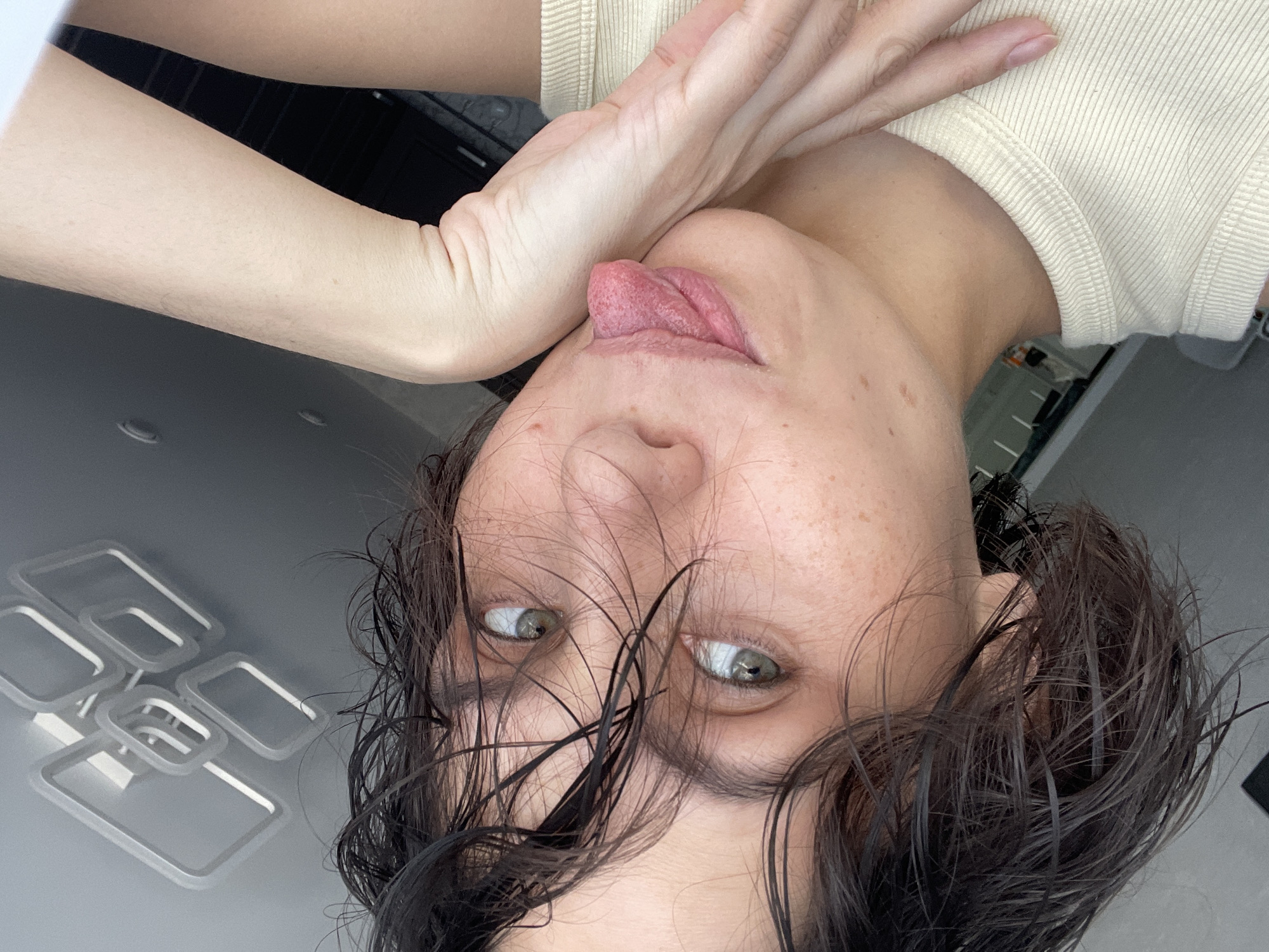 A woman with dark, wet hair is lying upside down, looking directly at the camera. She has fair skin with freckles and green eyes. Her tongue is sticking out, touching her hand, which is pressed against the side of her face. She is wearing a light-colored, short-sleeved shirt. The background includes a white ceiling fixture and what appears to be a glimpse of a dark interior. There is no indication of the location of Izhevsk, Russia, from this image.