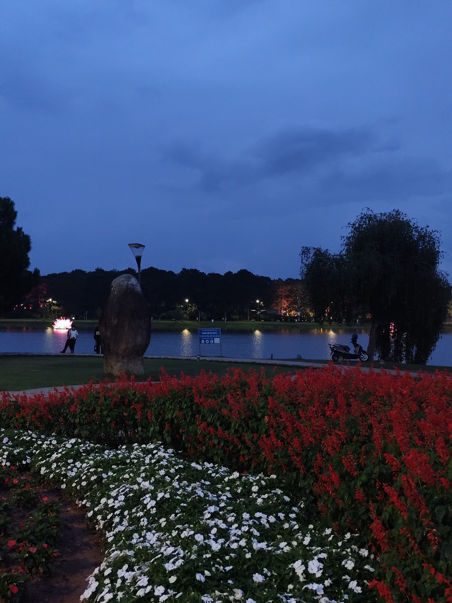 The image depicts a scene at Xuan Huong Lake in Da Lat, Vietnam at dusk. The foreground features a flower bed with red and white flowers. Behind the flowers is the lake. There is a rock with a light fixture on top of it along the side of the lake. People are walking in the distance. A motorcycle is parked under a tree, to the right. The sky is overcast and dark.