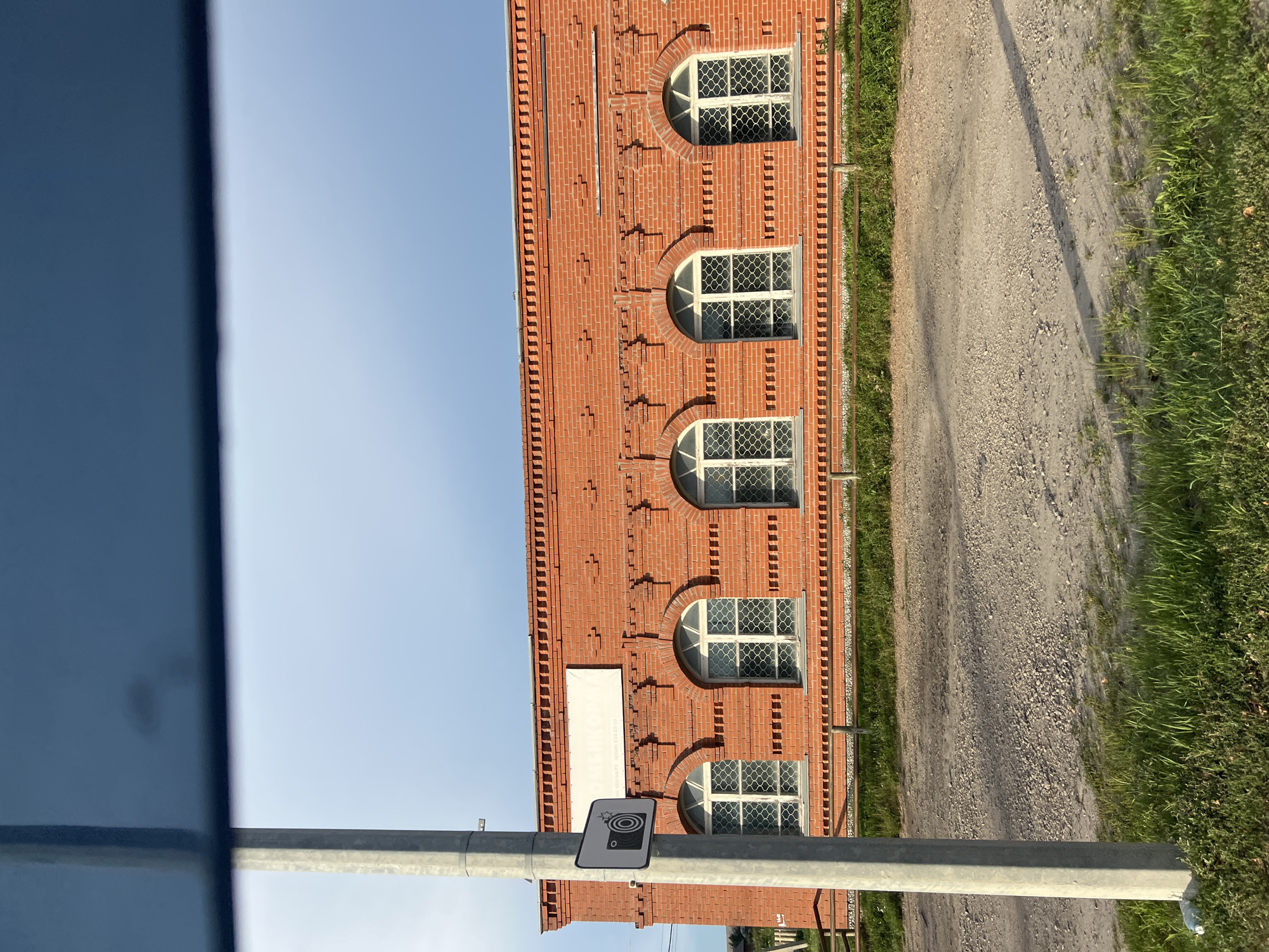 The image depicts a section of a red brick building with arched windows, located in the city of Bolshoy Oyosh, Russia. The building features decorative brickwork along the top and around the windows. A white rectangular sign is mounted on the facade above the windows. To the left of the building, a grey metal pole stands vertically, displaying a sign with a camera icon and radiating lines. A wooden fence runs along the base of the building. The foreground consists of a gravelly dirt road with patches of green grass on either side. The sky above is clear with a light blue hue.