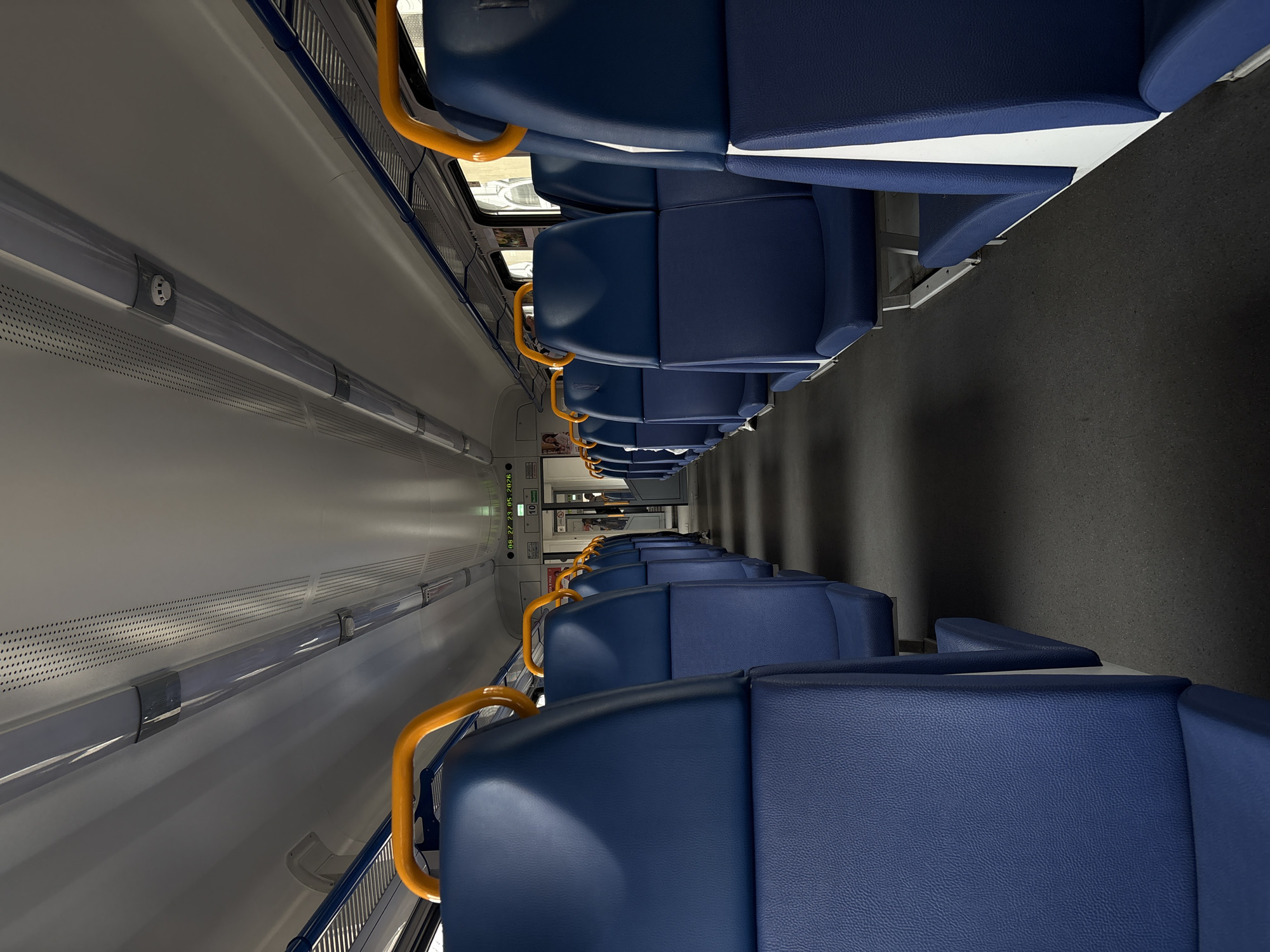 The image depicts the interior of a train car, featuring rows of blue seats with yellow handrails along the aisle. The walls are light gray, with ventilation panels and lighting fixtures running along the top. The floor is dark, likely carpeted. The train car is empty, with no visible passengers or staff. The perspective suggests the photo was taken from a high vantage point, possibly near the ceiling. The image does not provide any information about the location in the city of Khabarovsk, Russia.